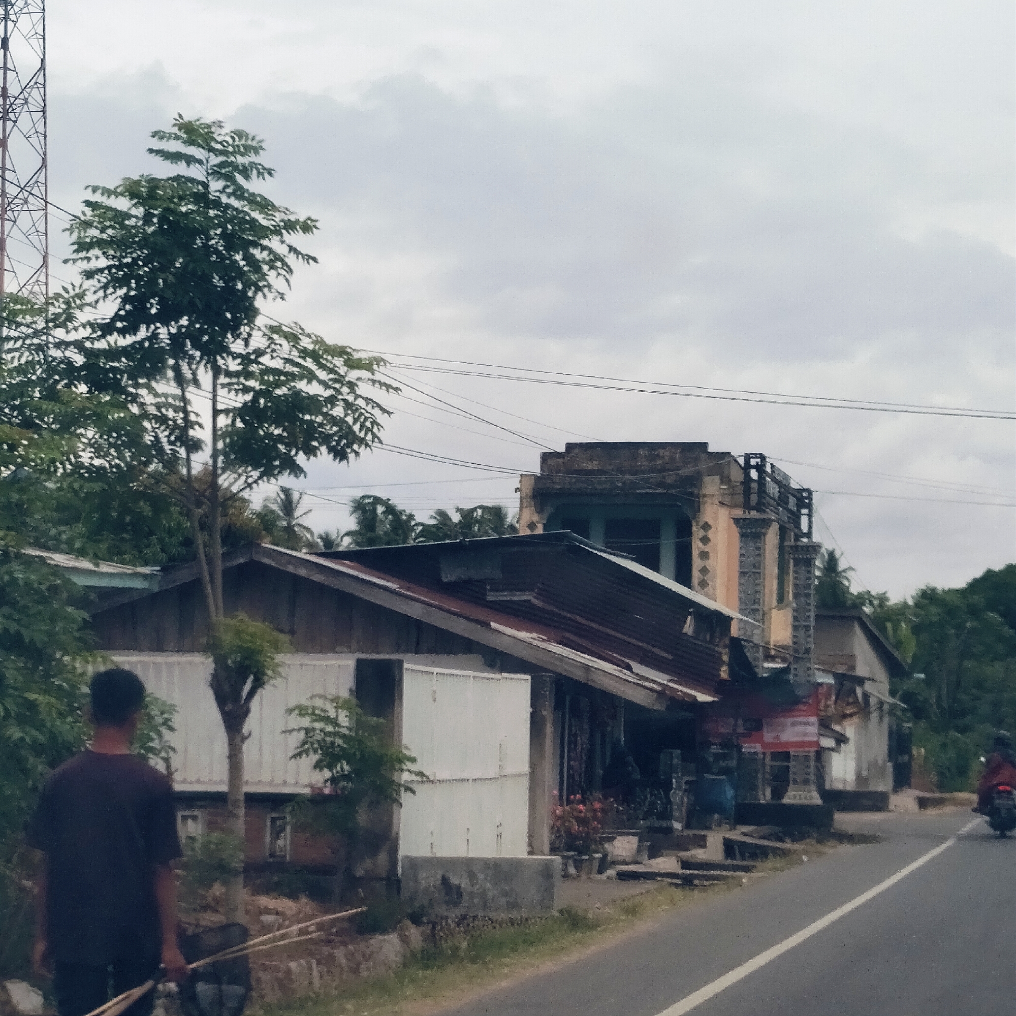 The image captures an overcast daytime street scene in Sawang, Indonesia. In the foreground on the left, a person in a dark t-shirt walks away from the camera, carrying long, thin sticks or poles. To their right is an asphalt road with a white solid line. Further down the road on the right, a person on a dark-colored motorcycle rides away from the viewer.
The right side of the street is lined with various buildings. Closest to the foreground is a single-story structure with a corrugated metal roof and white paneling. Further along the street stands a two-story building featuring an older, weathered facade with architectural elements like columns and a balcony railing on the upper floor. Other simpler buildings are visible in the background. Several trees with dense green foliage are prominent, including a tall tree in the mid-ground on the left, and palm trees are visible in the distance, characteristic of a tropical setting.
A large metal lattice tower, possibly for communication or utilities, is partially visible on the far left. Multiple power lines stretch across the grey, cloudy sky. Some shops appear to be present among the buildings, with indistinct items like speakers and blue containers visible outside one. Red and white signs are affixed to one building, but their text is illegible. The scene depicts a calm, typical day in a rural or semi-urban Indonesian street.