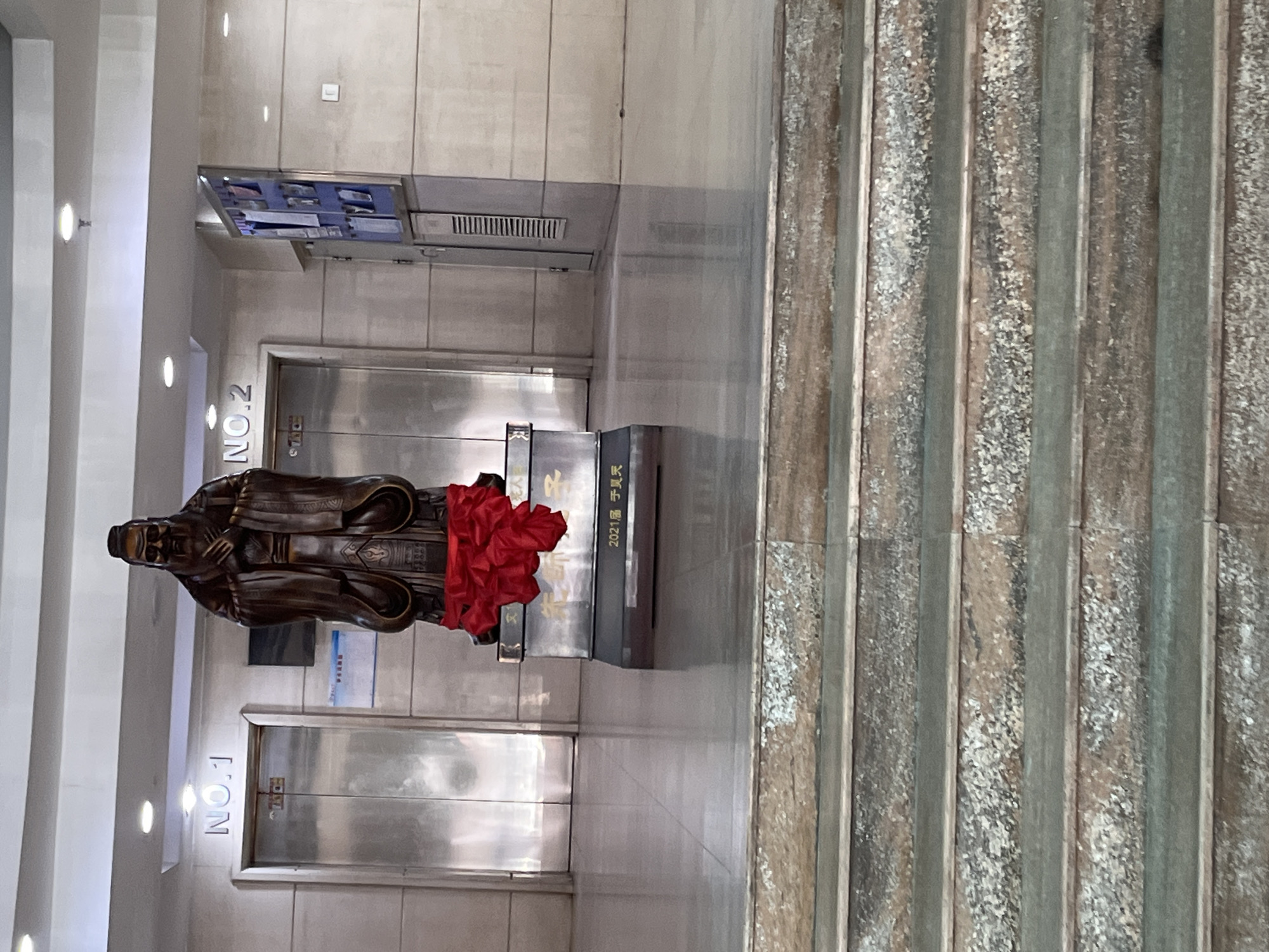 The image shows a statue of a man in a long robe, draped with red fabric, standing on a pedestal in a hallway. The pedestal has Chinese writing on it, including the year "2021". Behind the statue are two stainless steel doors labeled "NO.1" and "NO.2". The hallway has white walls with recessed lighting. To the right of the statue are textured, vertical panels. The location is in Changchun City, China, based on the Chinese writing and cultural context.