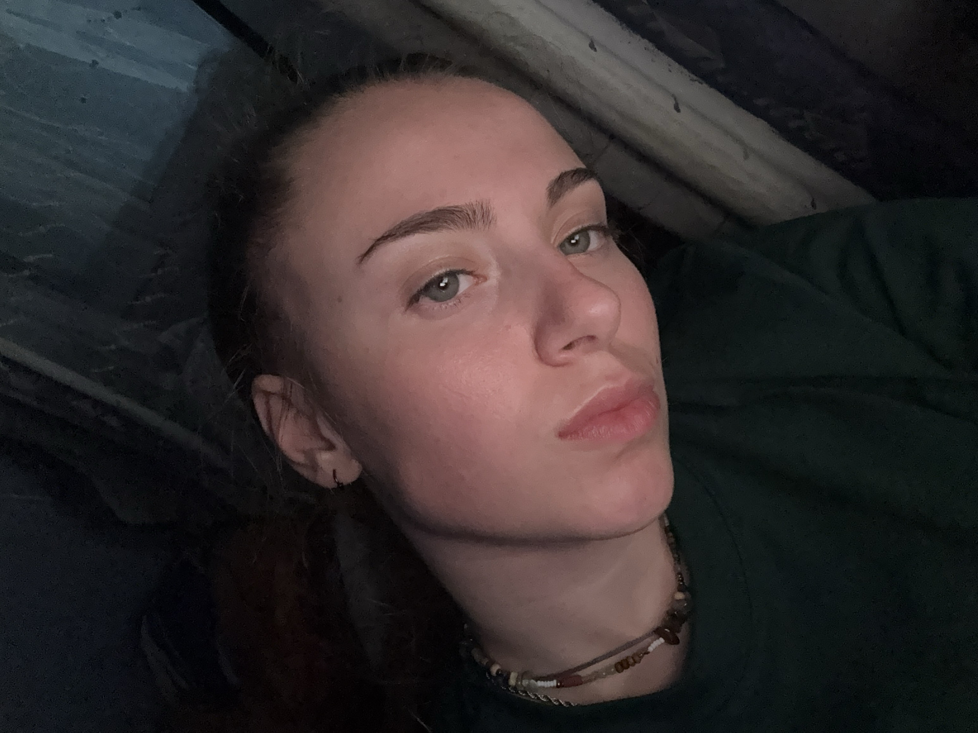 The image depicts a young individual, likely female, captured in a close-up portrait, with the frame rotated counter-clockwise. The scene is set in the city of Sofia, Bulgaria, within an indistinct, dimly lit indoor or enclosed environment.
The person has fair skin, light-colored eyes (appearing grey-green), and dark, well-defined eyebrows. Her hair, dark brown with reddish undertones, is pulled back from her face. She is wearing a dark green t-shirt. Around her neck, she has a multi-strand necklace composed of various small beads in earthy tones, including browns, white, and black. A small, dark stud or hoop earring is visible in her left ear. Her lips are naturally pink and slightly parted, contributing to a neutral facial expression. She looks directly towards the viewer.
The background is predominantly dark and lacks specific identifiers. On the right side of the frame, vertical, textured elements are discernible, resembling corrugated material, folded fabric, or possibly a tent wall, highlighted by ambient light from an unseen source to the upper left. The upper and left portions of the background are uniformly dark. The overall lighting is low and uneven, casting soft shadows.