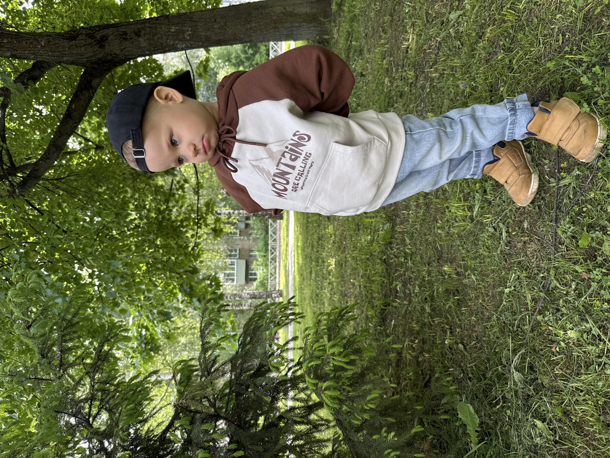 A young child wearing a two-toned brown and white hoodie with the word "MOUNTAINS" printed on it, blue jeans, and brown shoes with velcro straps is standing on a grassy lawn. The child is also wearing a black baseball cap and is looking down. The child is standing near a tree and a coniferous bush. In the background, a building with multiple windows is visible, and a white fence borders a grassy area. The location is in the city of Unknown, Unknown.