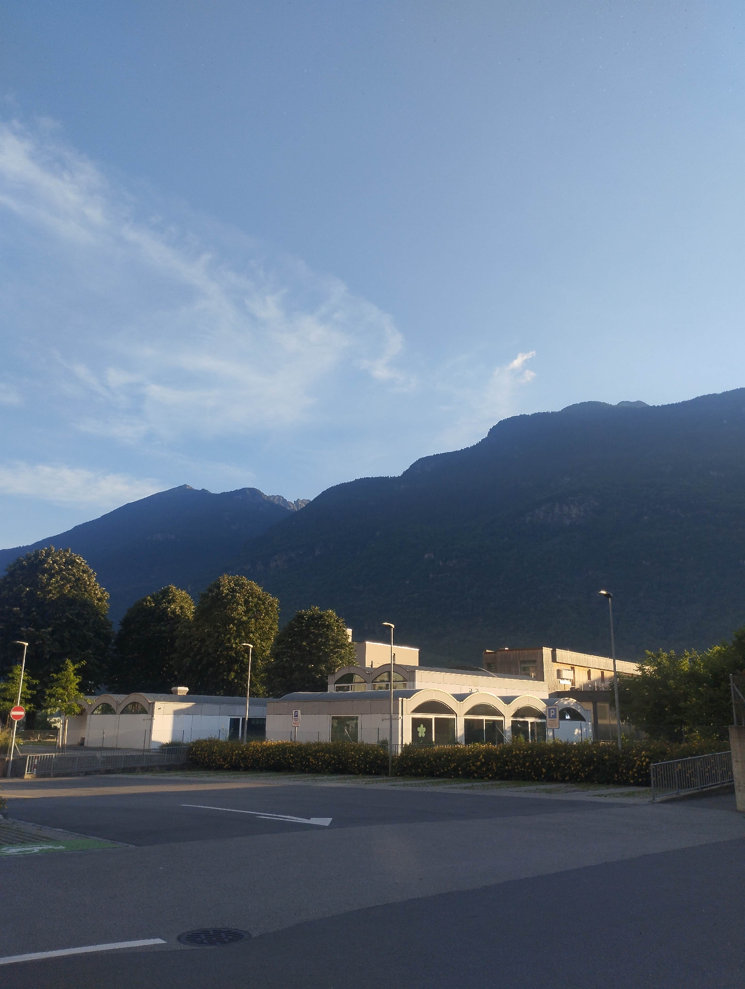 This forensic description details an outdoor scene in Biasca, Switzerland, captured on a clear day, likely during morning or late afternoon. The dominant background feature is a dark, heavily forested mountain range stretching across the entire upper half of the image, appearing deep green to black due to being in shadow. The sky above is a light, clear blue with wispy white cirrus clouds scattered across it, and a few small cumulus clouds near the mountain peaks.
In the midground, a collection of modern buildings is visible. The most prominent structure, centrally located, features a light gray facade with multiple distinctive arched window or entrance openings. To its right and slightly behind, a taller building with a light-colored, possibly wooden, exterior is present. A dense, low hedge with yellow flowering plants runs in front of these buildings, separating them from a paved area. Numerous mature, deciduous trees with full green canopies are interspersed around the buildings and extend towards the base of the mountains, with several large trees prominently silhouetted on the left.
The foreground consists of an asphalt road and paved areas. A white arrow is painted on the road, pointing diagonally right and upwards, indicating a directional turn or entrance. A circular metal manhole cover is embedded in the lower center of the road. On the far left, a green painted strip with a partial white bicycle symbol is visible, suggesting a bicycle lane. Several tall, slender streetlights with modern fixtures illuminate the scene. Street furniture includes a red circular "No Entry" sign mounted on a pole on the far left and a blue rectangular "P" parking sign on a pole near the central building. A metal railing delineates a ramp or elevated section on the right side of the paved area. No individuals or vehicles are present in the scene. The overall lighting suggests the sun is low in the sky, illuminating the foreground buildings directly while casting the mountains into shadow.