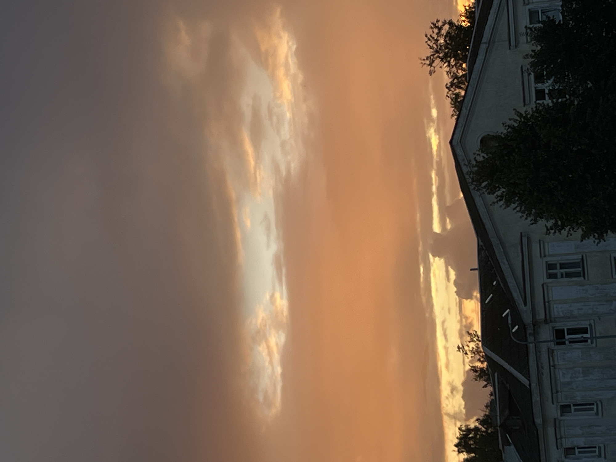 The image depicts a cloudy sky over a building, likely in Kohtla-Järve linn, Estonia. The sky is dominated by a gradient of colors, transitioning from dark gray at the top to a vibrant orange and yellow near the horizon, indicating a sunset or sunrise. Wispy clouds are scattered across the sky, with some catching the light and appearing brighter.
Below the sky, the upper portion of a building is visible. It appears to be a multi-story structure with a dark, possibly tiled, roof. The building's facade is light-colored, with visible windows and architectural details. Some trees are silhouetted against the sky and the building, suggesting they are located in front of or around the structure. The overall impression is one of an urban landscape bathed in the warm light of dusk or dawn.