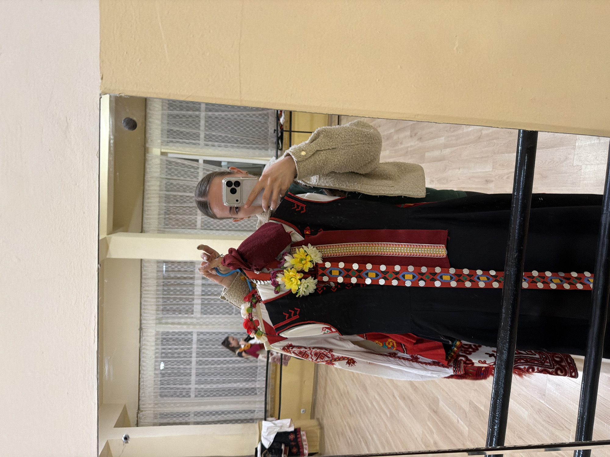 An indoor mirror selfie captures a young woman holding several traditional garments, likely Bulgarian folk costumes. The image is taken in what appears to be a studio or rehearsal space, characterized by light-colored walls and a wooden floor visible in the reflection.
The central subject, a woman with dark hair pulled back, holds a silver iPhone with a clear case and three camera lenses, taking the picture. She is wearing a light beige, textured (possibly sherpa-style) jacket over a dark green top. Her arm extends to hold a hanger from which several elaborate costumes are suspended.
The prominent costume is a dark, possibly black, garment featuring intricate embroidery and embellishments. It has a broad red band with geometric patterns in blue, yellow, and white, adorned with circular, silver-colored metallic elements resembling coins or sequins. Yellow and white artificial flowers are pinned or sewn onto this garment. Beneath this, parts of other costumes are visible, including a white garment with red and black embroidered patterns.
In the reflection behind the woman, windows with horizontal blinds are visible, suggesting an external light source or view. Another blurry figure is faintly reflected further back in the room. No discernible text is visible in the image. The costumes strongly suggest a cultural or performance context, aligning with the location context of Sofia, Bulgaria.