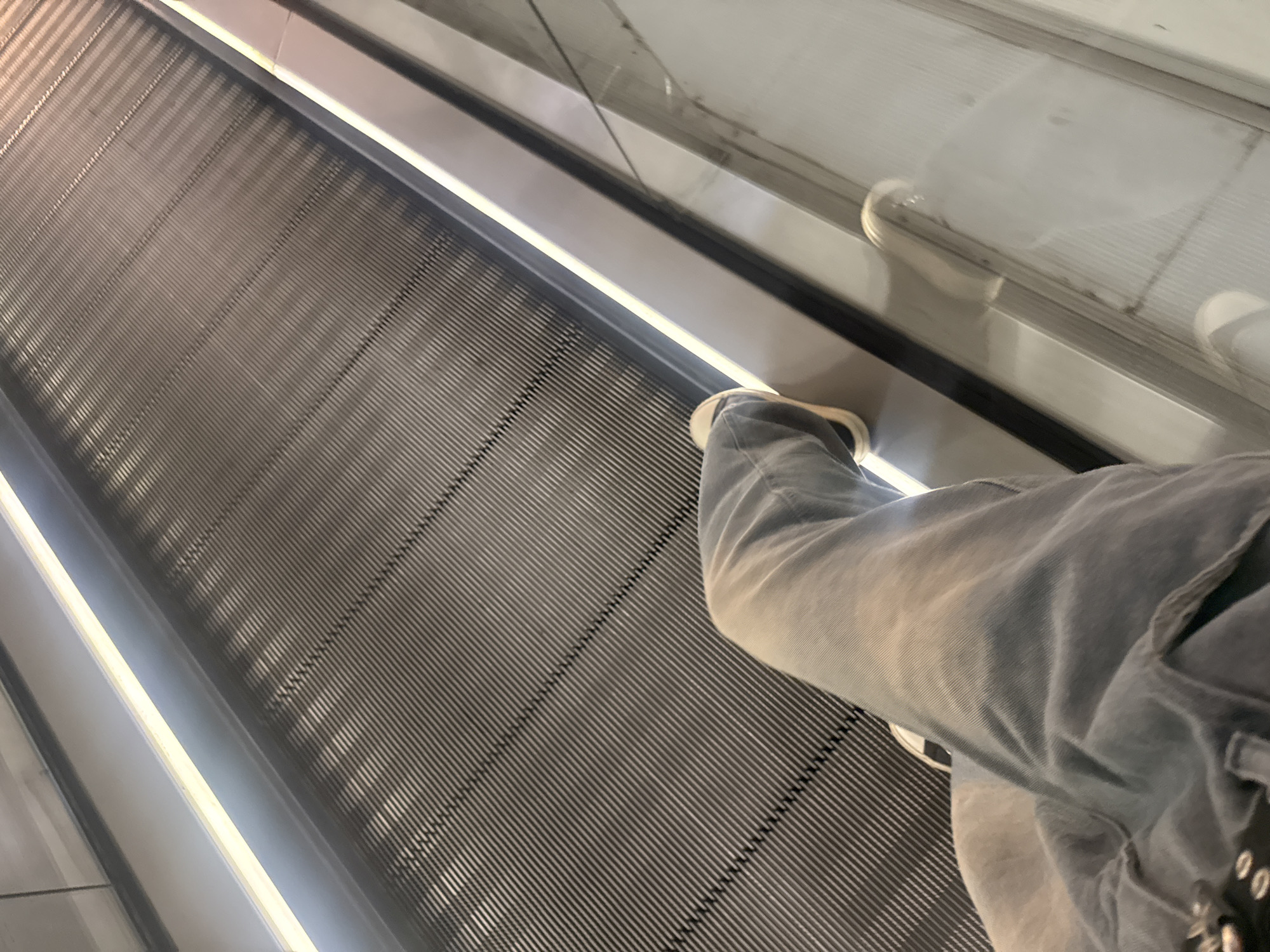 A first-person perspective shot of someone riding an escalator. The person is wearing grey jeans and white shoes. The escalator is grey with dark lines, and the sides are lined with bright lights. The location appears to be an indoor space. It is not possible to determine the location in Saint Petersburg, Russia from this image.