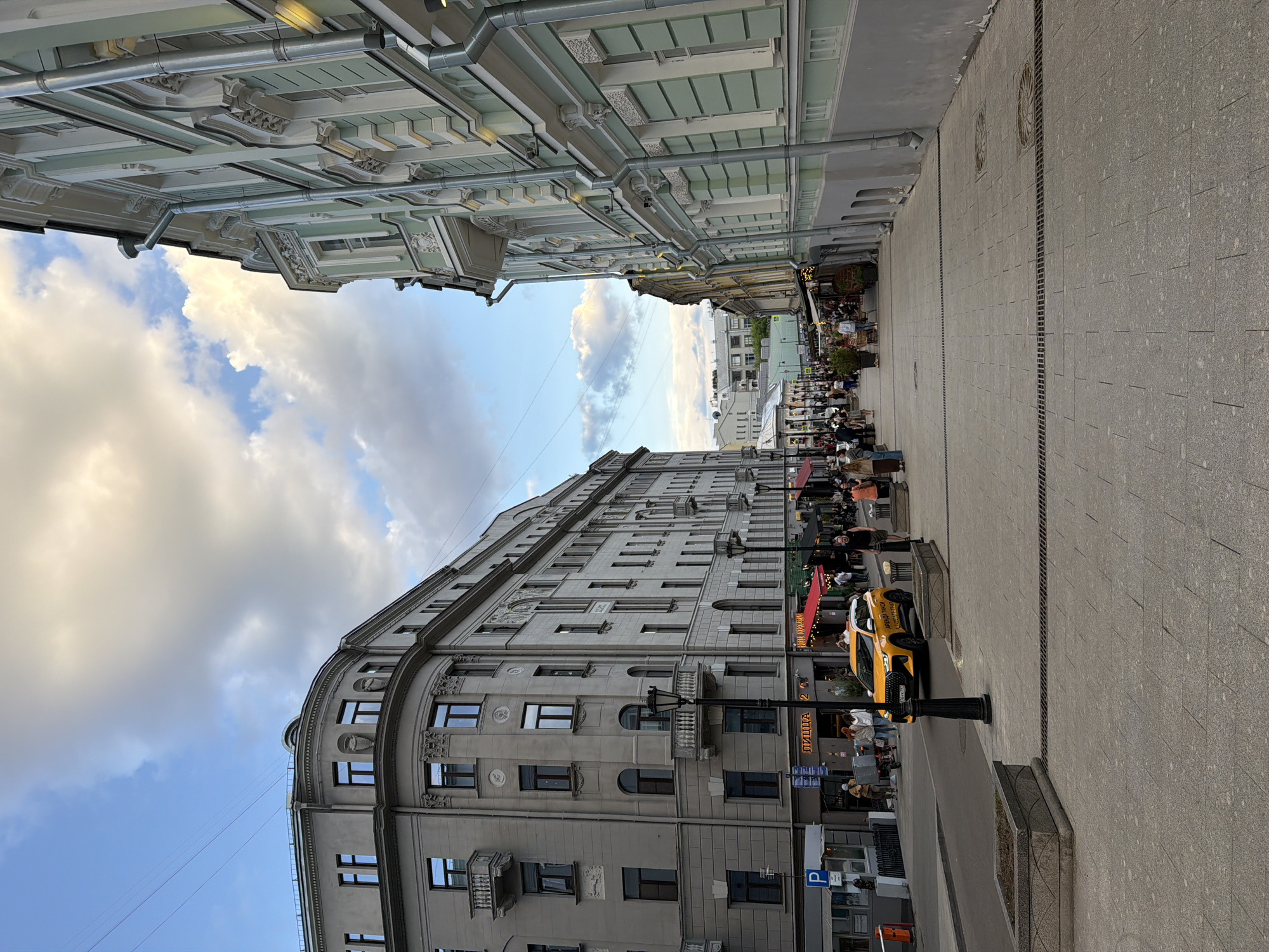 The image depicts a street scene in an urban environment, likely during the day. The camera is positioned at street level, looking down a pedestrianized thoroughfare.
Two buildings flank the street: one on the left is taller and clad in a neutral gray tone, while the other has a lighter color and a more ornate architectural design. The sky is visible between the buildings, revealing a mix of blue and cloudy patches.
People are present along the street, some seated at outdoor tables, suggesting a restaurant or cafe setting. A yellow car, possibly a taxi, is parked on the right side of the street. Streetlights and other urban fixtures are also visible along the walkway.
The street surface is paved with stone or concrete tiles, featuring parallel drainage lines. The overall ambiance suggests a bustling urban scene with elements of historical architecture.
The location is Unknown, Unknown.