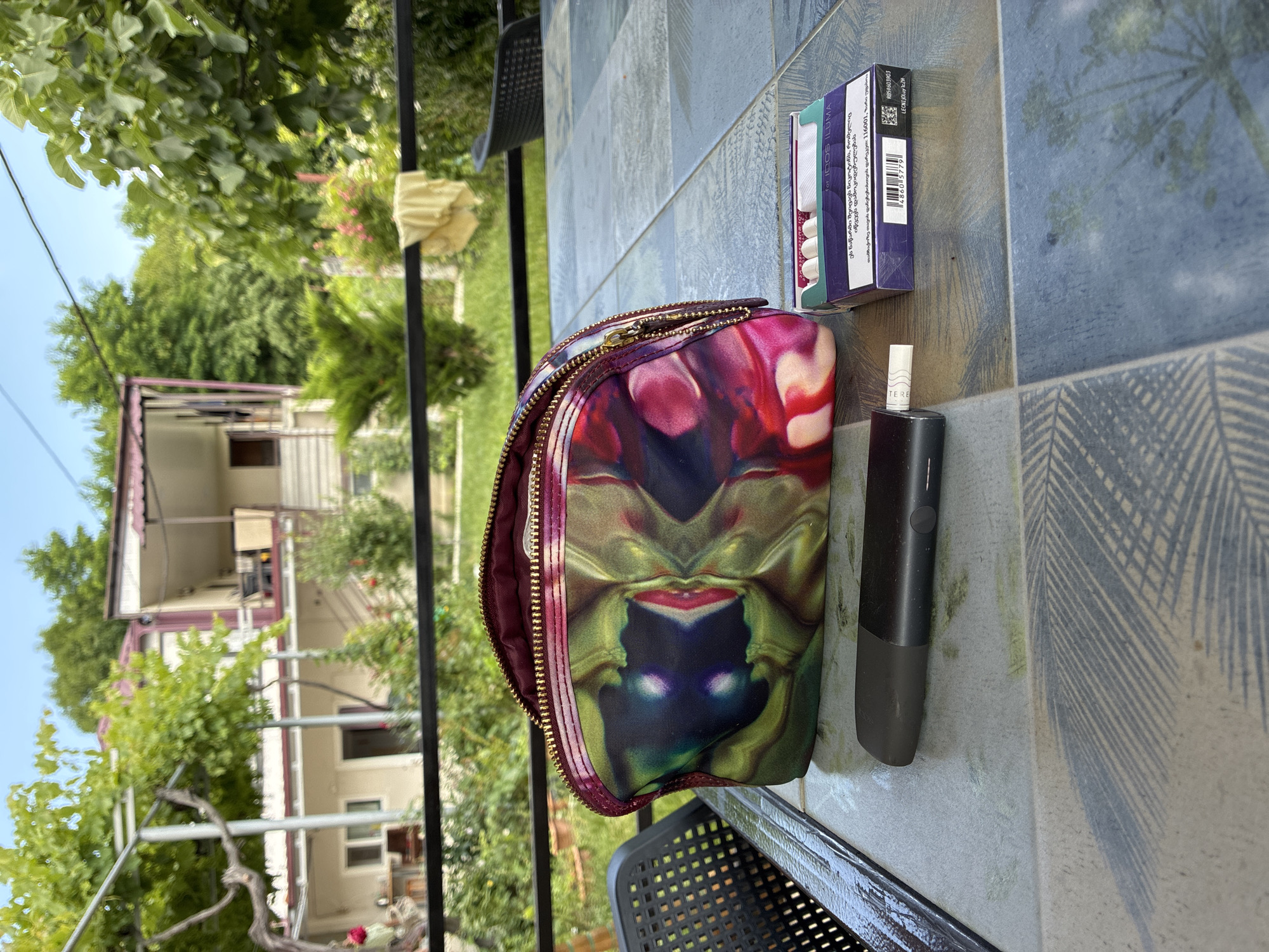 A close-up, eye-level shot captures a table with several items, suggesting a casual outdoor setting. The table's surface features a patterned mosaic of blue, grey, and white tiles with abstract designs. In the foreground, a black electronic smoking device rests horizontally, with a white filter tip partially inserted into its end. To the right of the device is a purple and white box of tobacco sticks, identified as "for IQOS ILUMA" by visible text. A vibrant, multi-colored makeup bag, adorned with a psychedelic pattern of greens, purples, and reds, is positioned behind the smoking device and tobacco. The bag's zipper is partially open, revealing its interior.
The background suggests a residential area in Surami, Georgia, with a two-story house visible behind a black metal railing. The house has a light beige exterior with a maroon roof and balconies. Lush green foliage, including trees and vines, surrounds the house and extends across the backdrop, indicating a temperate climate. The overall impression is one of a relaxed, domestic scene. The lighting indicates it is daytime.