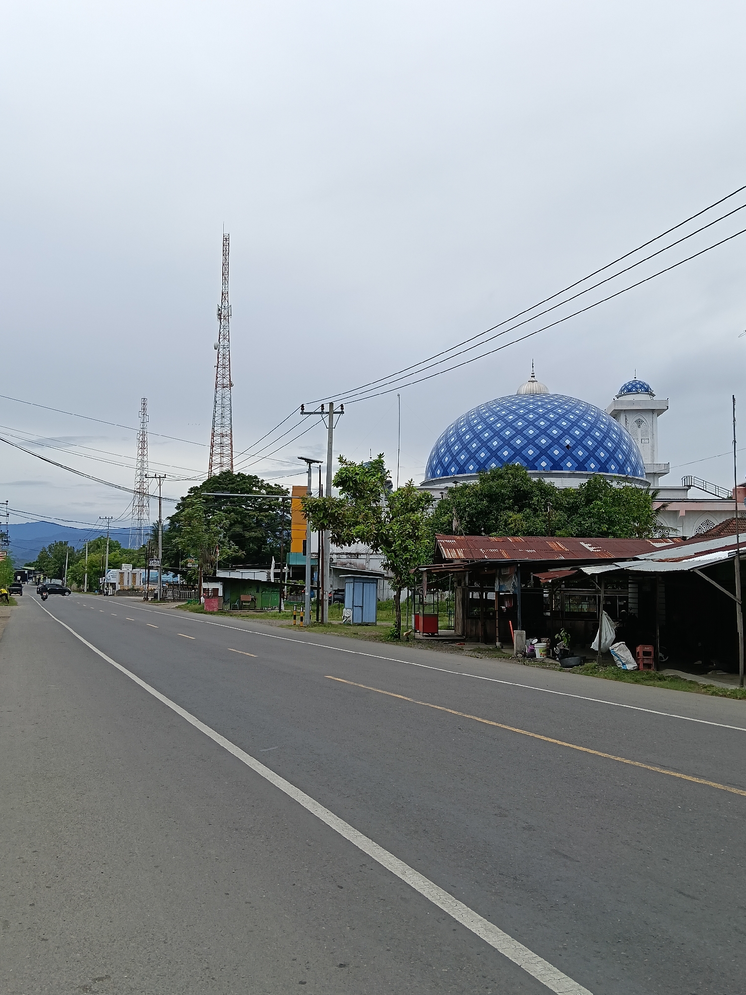 The image depicts a street scene in Paya Tringgadeng, Indonesia. A two-lane asphalt road runs diagonally from the lower left to the upper right of the frame, with a white solid line on the left edge and a broken yellow line in the center. On the right side of the road, there are several structures, including a prominent mosque with a large blue and white checkered dome and a smaller dome with a minaret. In front of the mosque, there are some low-lying structures with corrugated tin roofs, possibly market stalls or small shops. Various trees and foliage are present throughout the scene. In the background, there are communication towers, utility poles with wires, and a distant mountainous landscape under an overcast sky. A few vehicles, including cars and a motorcycle, are visible on the road, and some bags and containers are scattered near the roadside structures. The overall impression is one of a roadside view in a semi-urban or rural Indonesian setting.