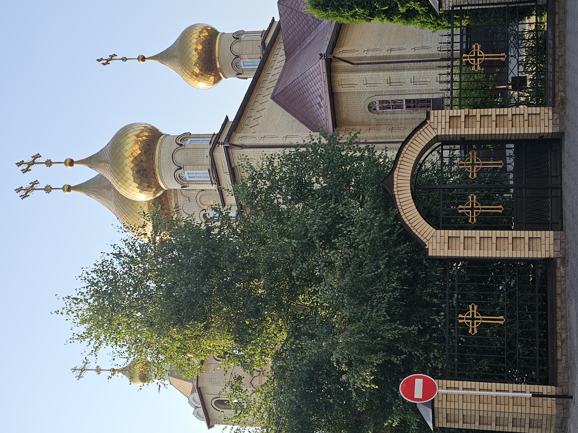 The image depicts the Church of the Archangel Michael in Cherkessk, Russia. The church is a yellow brick building with a copper-colored roof and multiple golden domes topped with crosses. In the foreground, a black metal fence with arched gates is adorned with golden crosses. A red and white "no entry" sign stands to the left of the fence. Lush green trees partially obscure the church building. The sky is clear and blue.