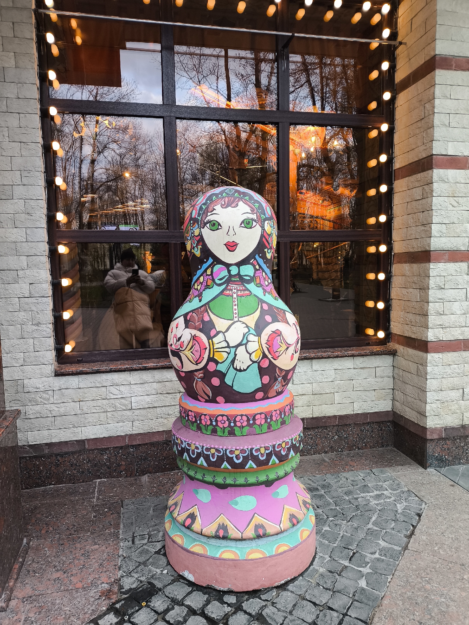 A brightly painted, very large matryoshka doll stands on a cobblestone surface adjacent to a building in the city of Unknown, Unknown. The doll is composed of stacked, disc-shaped segments that form its base, all adorned with geometric and floral patterns in pinks, greens, and oranges. The doll itself features traditional Russian doll characteristics like a round face with green eyes, rosy cheeks, and a red bow. It has a patterned headscarf, and its body is decorated with floral designs and other decorative elements in a range of colors. Behind the doll is a large, multi-pane window reflecting bare trees and a person with a phone taking a photograph of the doll. A string of lights hangs vertically along the building's brick façade on either side of the window. The building's wall is constructed of light-colored stone bricks, with a horizontal band of reddish-brown stone along the base. The cobblestone surface extends from the base of the doll towards the bottom of the image.