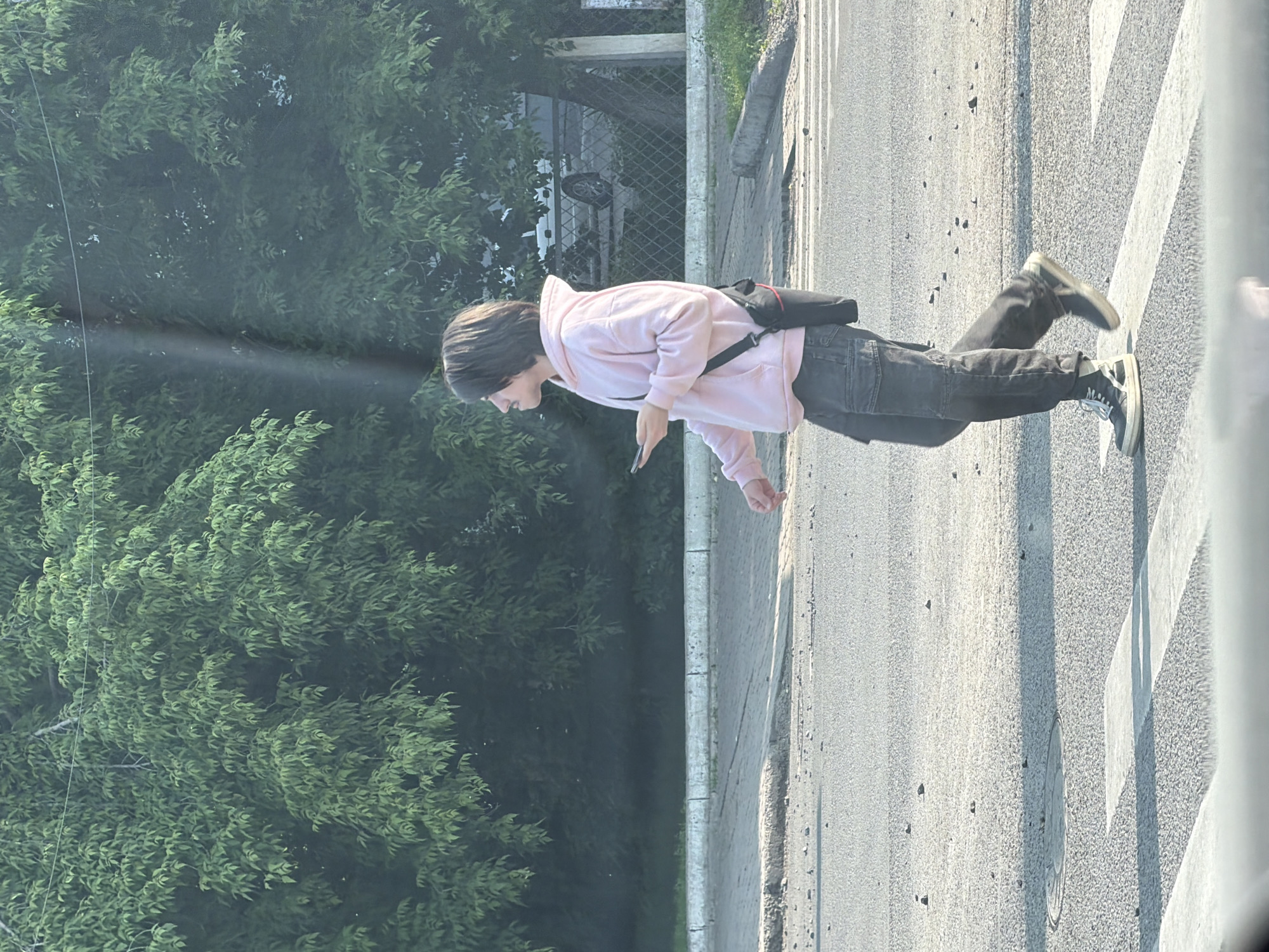 A young person with medium-length dark hair is walking across a street at a pedestrian crossing. They are wearing a light pink hooded sweatshirt, dark cargo pants, and black and white high-top sneakers. They are holding a cell phone in their right hand and have a black bag with a red strap slung across their chest. The street appears to be asphalt with scattered debris. In the background, there are trees with dense green foliage, a sidewalk, a curb, a chain-link fence, and a parked vehicle. The lighting suggests it is daytime, possibly late afternoon, with the sun casting shadows. This scene is located in the city of Krasnoyarsk, Russia.