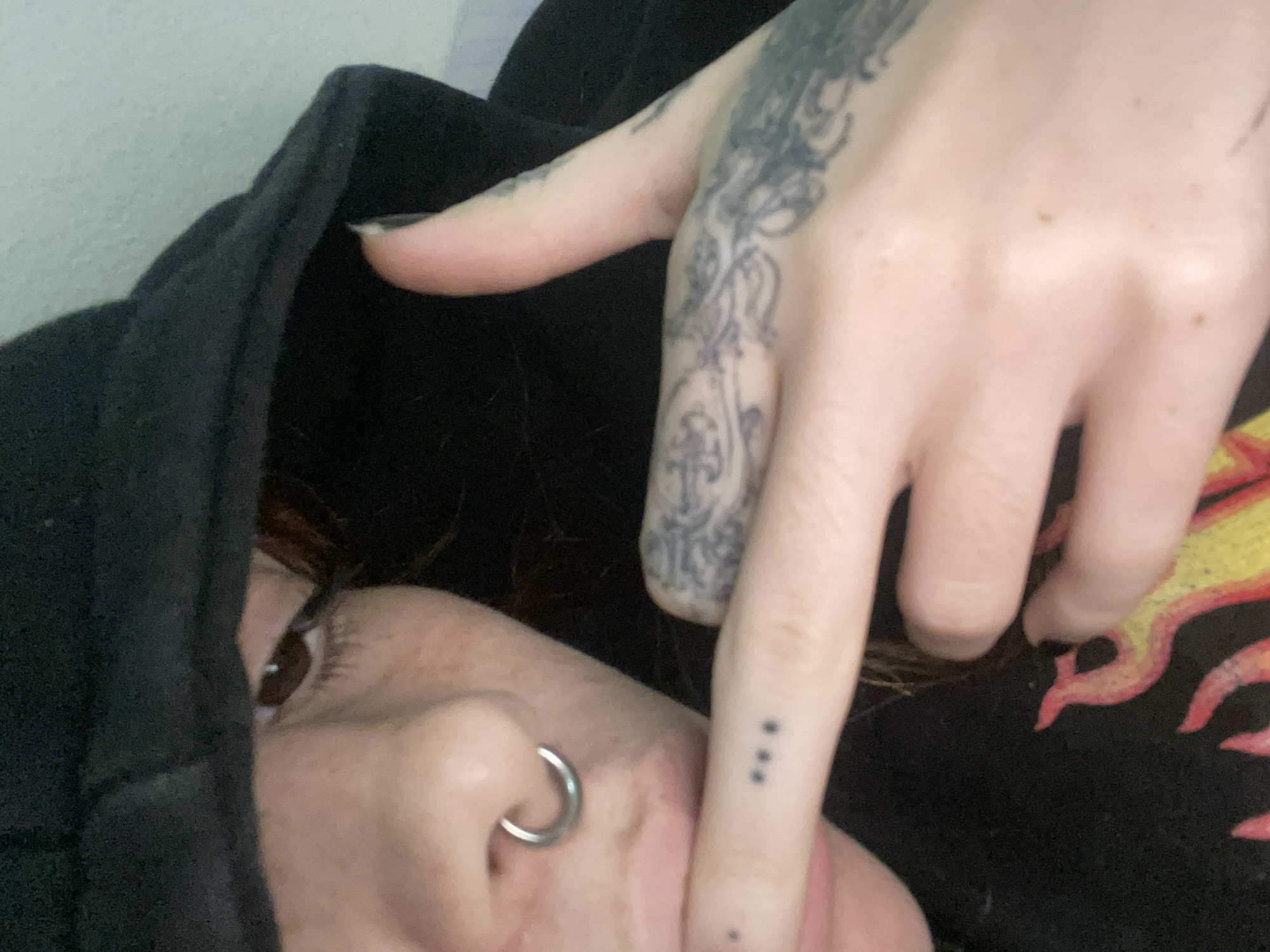 A close-up photograph shows a person partially obscured by a black hooded sweatshirt. The person has fair skin, freckles, a silver nose ring, and dark hair. Their right eye is visible. Their right hand is positioned in front of their face, with visible tattoos on the back of their hand and three small dots tattooed on their finger. The person's other arm and yellow and red flaming pattern material is on the other side of the picture behind their hand. The background appears to be a plain white wall. The image provides no information about the location in Buenos Aires, Argentina.