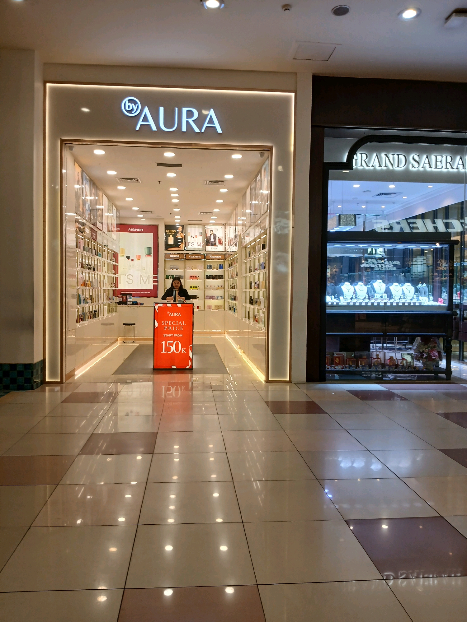 The image shows the entrance to a store called "AURA" in a mall in North Jakarta, Indonesia. The store appears to sell perfumes and cosmetics, with shelves lined with bottles visible through the glass front. A woman is standing behind a counter with a sign that advertises a "special price" starting from "150k". To the right of the AURA store is another store with a sign that reads "GRAND SAERA", which appears to be a jewelry store, displaying necklaces and earrings in a glass case. The floor of the mall is tiled in a checkered pattern of light and dark brown. The lighting in the mall is bright, with overhead lights reflecting off the shiny floor.