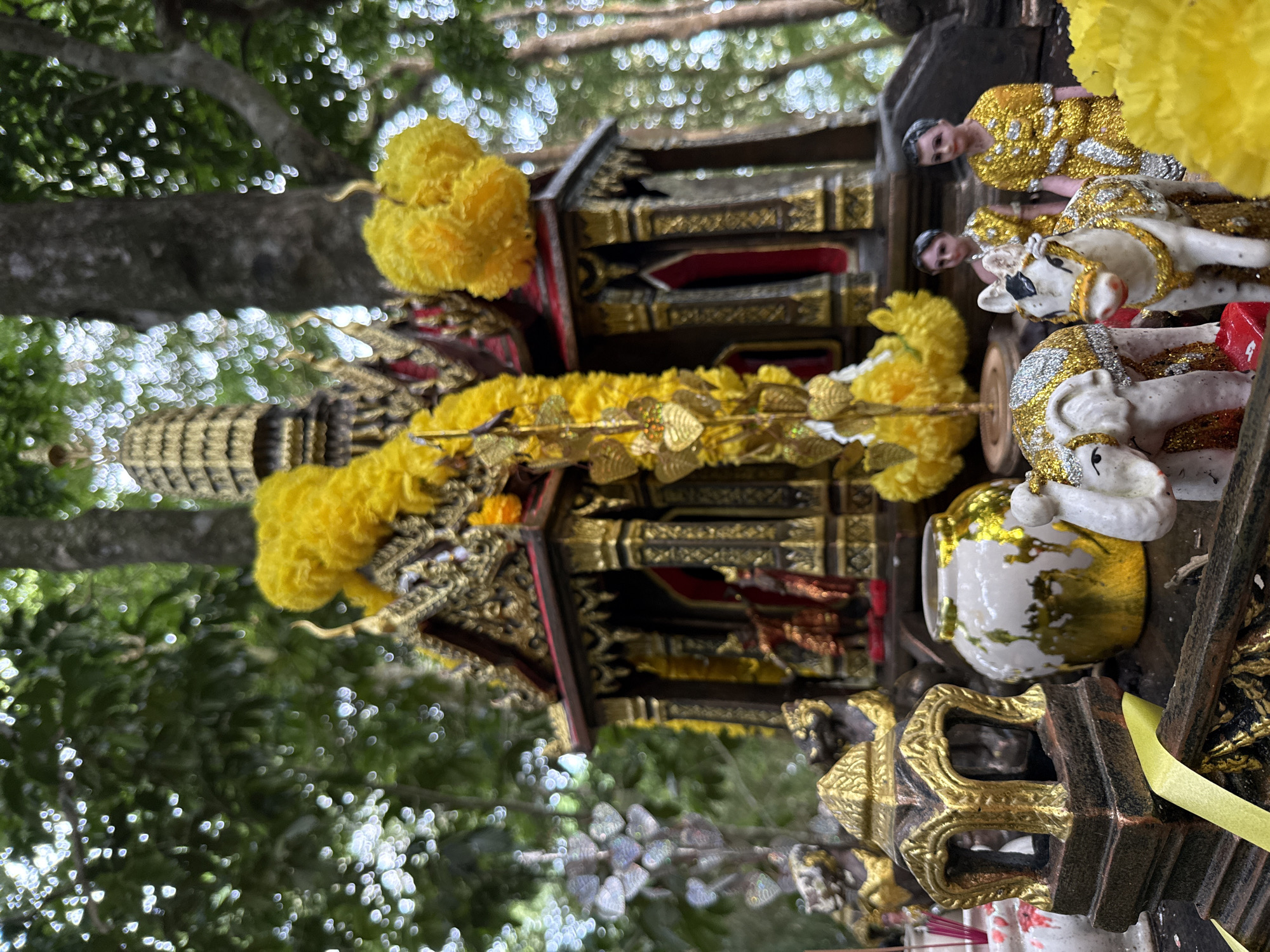 The image depicts an outdoor shrine, likely a spirit house, set within a natural environment with trees and foliage in the background. The shrine itself is ornate, with gilded architectural details and dark wood elements. It is decorated with vibrant yellow garlands and fluffy yellow pom-poms. Inside the shrine, two small figures, possibly deities or representations, can be seen. In the foreground, a collection of miniature figures and offerings are arranged. These include a white elephant figure adorned with glitter, a white horse figure, a patterned ceramic vase, and a small golden lantern-like object. The overall impression is one of a religious or spiritual offering site, possibly located in the vicinity of Ko Samui District, Thailand, given the common practice of such shrines in the region.