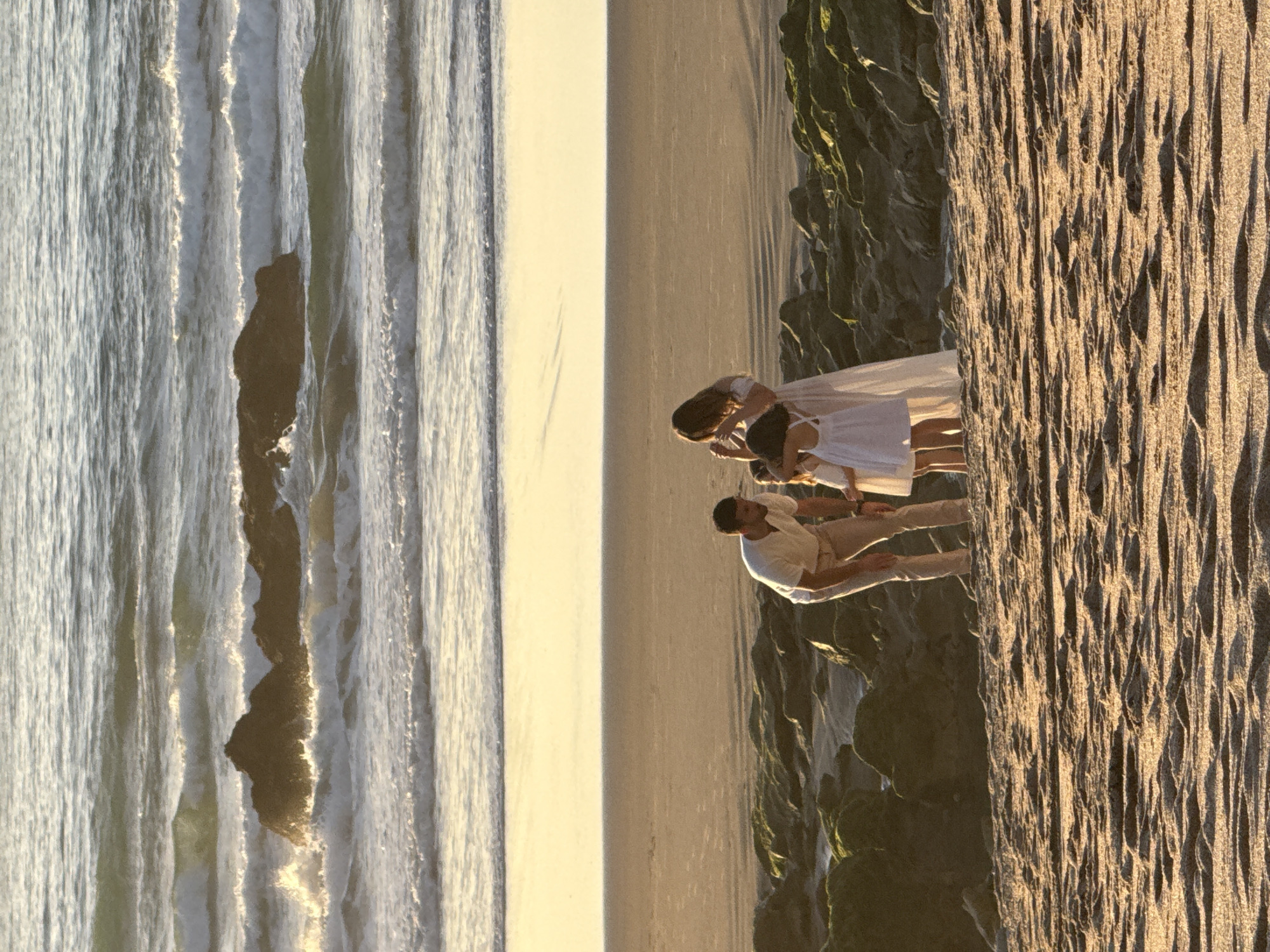 The image shows a family of four on a beach in Biarritz, France. The family consists of a man, a woman, and two young girls. The man is wearing a white t-shirt and beige pants, and is bending down to interact with the girls. The woman is wearing a white dress and is standing next to the girls. The two girls are wearing white dresses. They are standing on a sandy beach, with rocks and vegetation to their left. In the background, there is the ocean with waves crashing on the shore. The sun is setting, casting a warm glow over the scene.