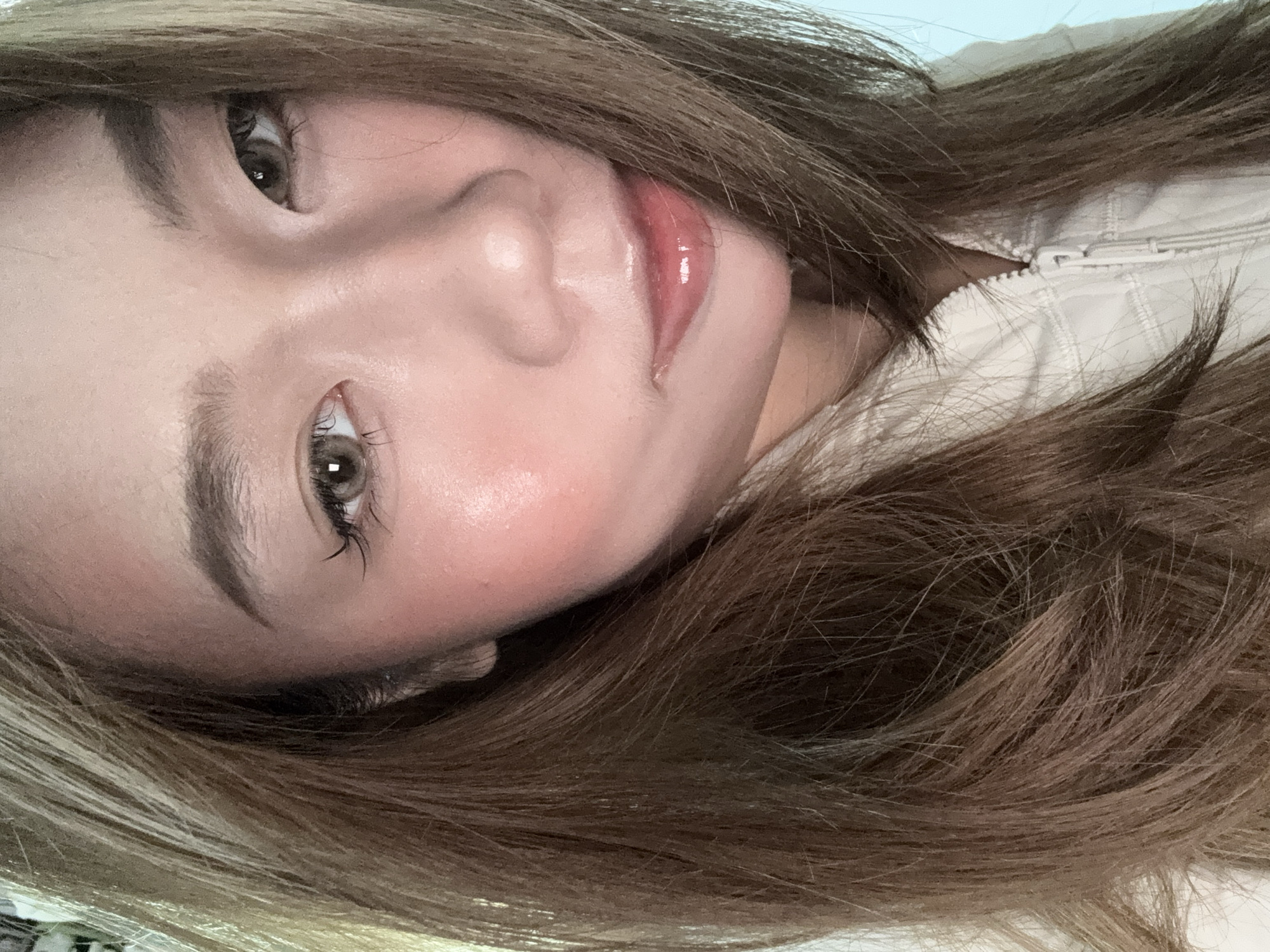 A close-up, rotated image shows a person with light brown hair, fair skin, and hazel eyes. The person is wearing lip gloss. A white zippered garment is visible at the upper right. The location is unknown, and no information suggests a location in Nan'an District, China.