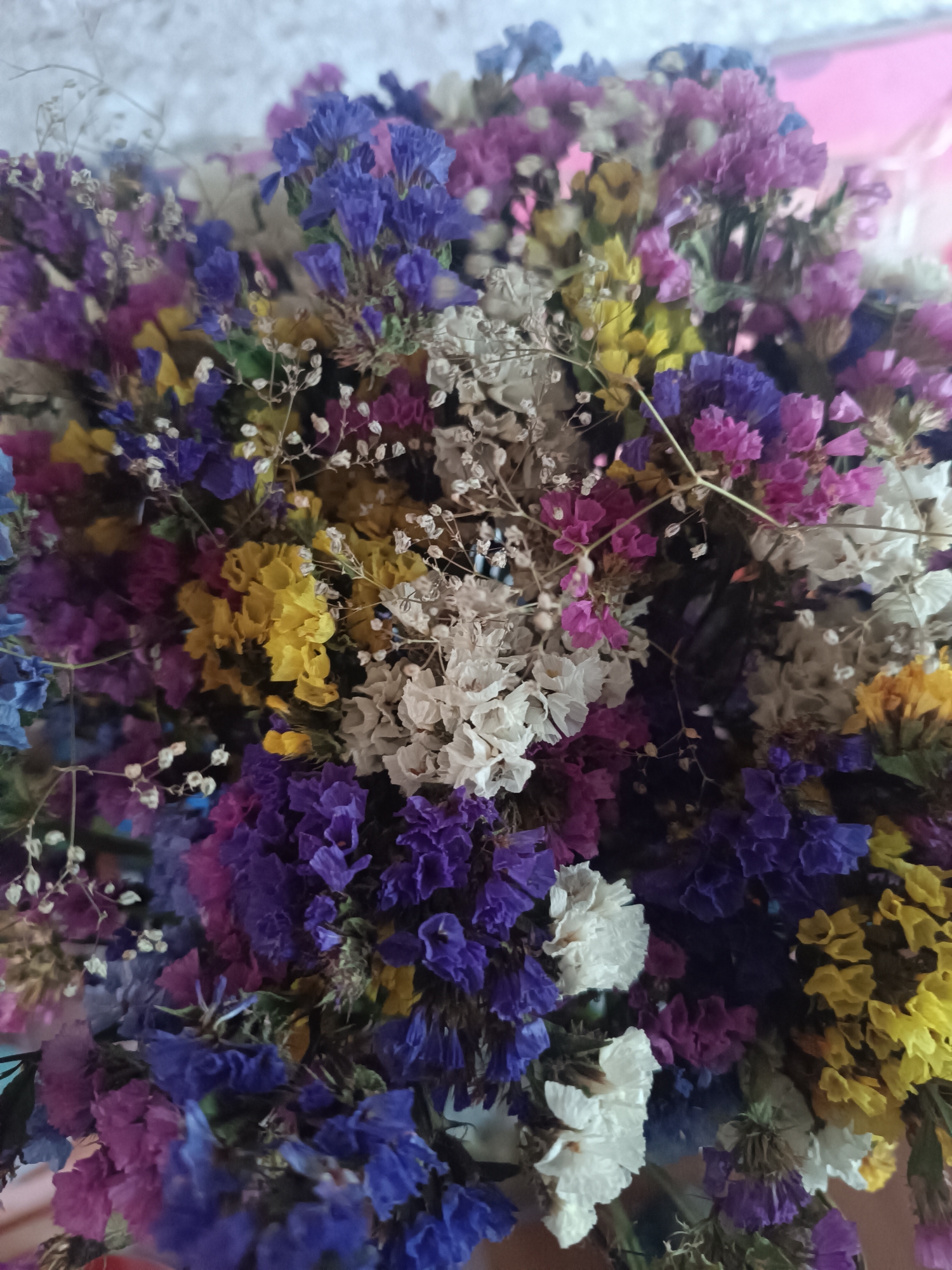 This is a close-up, eye-level shot of a bouquet of dried flowers. The flowers are small and clustered together, with various colors including purple, blue, yellow, white, and pink. Some of the flowers have delicate, feathery stems. The background is blurred and appears to be a light-colored, textured surface, possibly a wall. There are no people or animals visible in the image, and no specific location in Vypasne, Ukraine can be identified. The lighting is soft and diffused, creating a gentle overall mood.