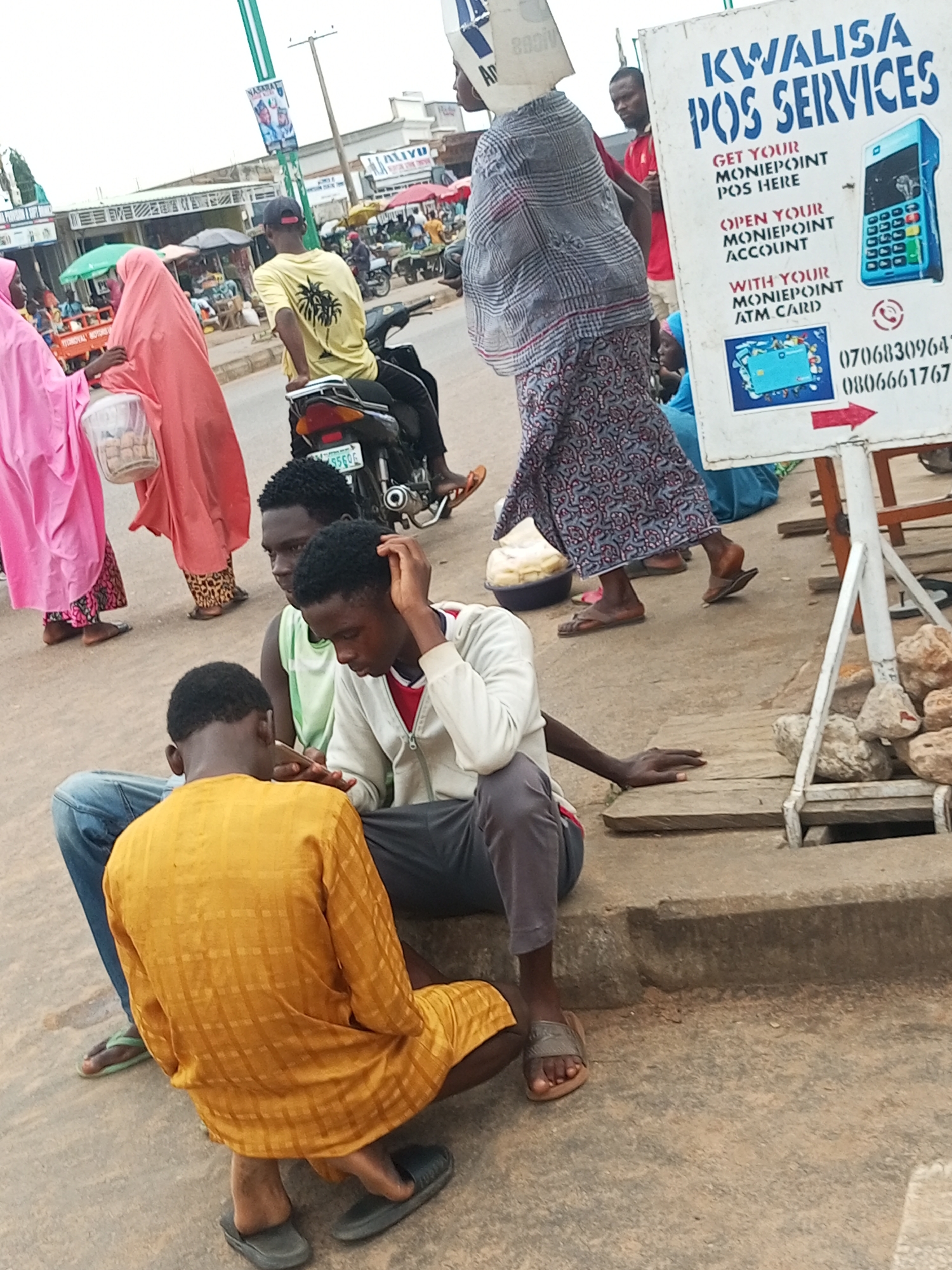 In an outdoor, urban environment, likely a marketplace, three young men are seated on a concrete curb. The man on the right wears a white hooded jacket over a red shirt and dark pants. He is looking down at a smartphone in his hand. The man in the middle, wearing a light green sleeveless shirt, is also looking at the phone. The man on the left is wearing a yellow checkered tunic and blue jeans, and he is facing away from the camera, looking down at the phone as well. A motorcycle with a partially visible license plate is parked behind them. In the background, several people are walking, some wearing traditional clothing. A large white sign with blue text advertises "KWAIISA POS SERVICES." Other stalls and vendors are visible in the distance, along with street poles and buildings. The ground is paved with concrete and some dirt. The lighting suggests it is daytime.