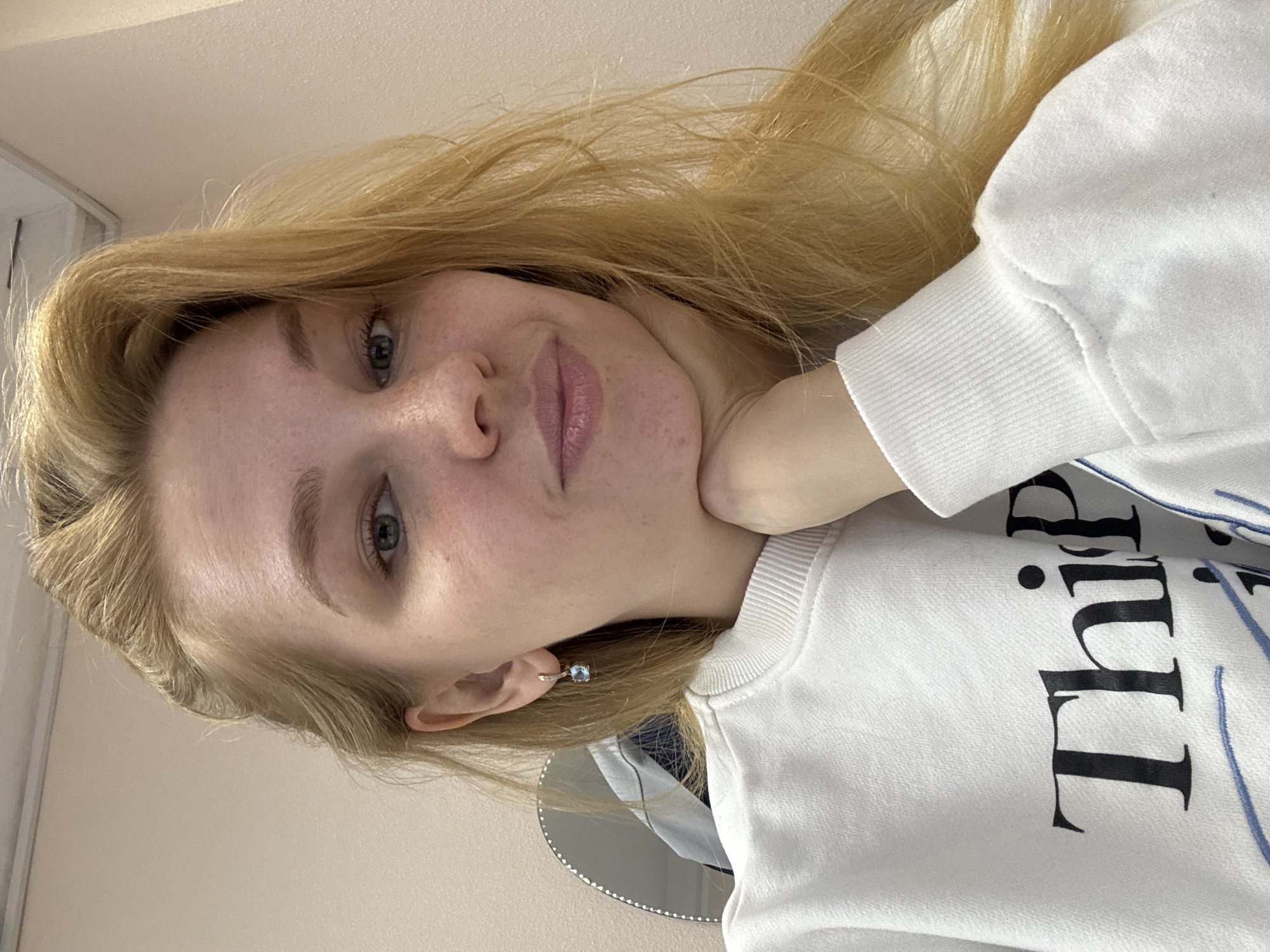 A fair-skinned young woman with blonde hair is posing for a selfie. She is wearing a white sweatshirt with black lettering and blue accents. She is wearing a small stud earring with a blue stone. The background is a light beige wall. The location cannot be determined as Rusanovo, Russia without additional information.