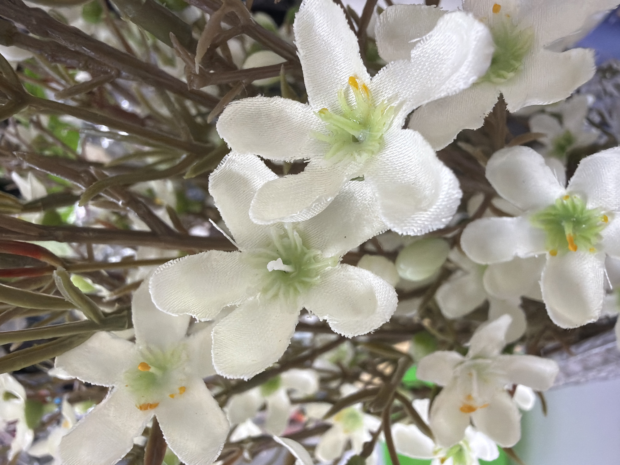 The image presents a close-up, macro-style view of an artificial floral arrangement. The primary subjects are multiple white, five-petaled flowers. Each flower is constructed from a textured fabric material, possibly velvet or a similar textile, exhibiting a soft, slightly napped surface and minor irregularities along the petal edges. The center of each blossom features a distinct light green, upright pistil-like structure, surrounded by several small, bright yellow, granular anthers or stamens, typically 3 to 5 per flower. These flowers are attached to slender, branching stems that are dark brown to olive-green in color and appear to be artificial, likely made of plastic-coated wire or molded plastic. Some stems exhibit rudimentary leaf or bud forms. The arrangement is densely packed, with numerous flowers and stems occupying the frame. The background is heavily blurred and indistinct, showing soft transitions between white, various shades of green, and some darker tones, suggesting an indoor environment or a display setting. No individuals or specific objects are discernible in the background. The visual content of the image does not provide any identifying characteristics or contextual cues to determine a specific location within the city of Sochi, Russia, or any other geographic area.