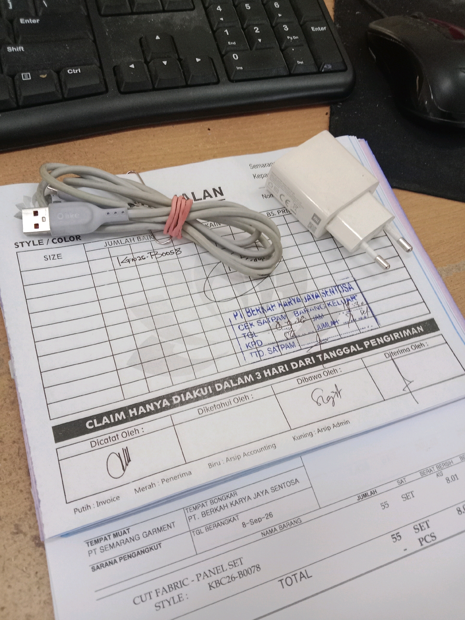 A flat lay image shows a paper form, a white USB charger, and a gray USB cable on a wooden surface. The form appears to be a shipping or receiving document. Handwritten text in Indonesian indicates information about the sender, recipient, and items. Some of the text suggests it is related to a company in Semarang, Indonesia, specifically "PT. Berkah Karya Jaya Sentosa" located in "Semarang." The charger has the "MI" logo and "CE" marking. The cable has "Olike" branding. The scene is likely an office or workspace environment.
