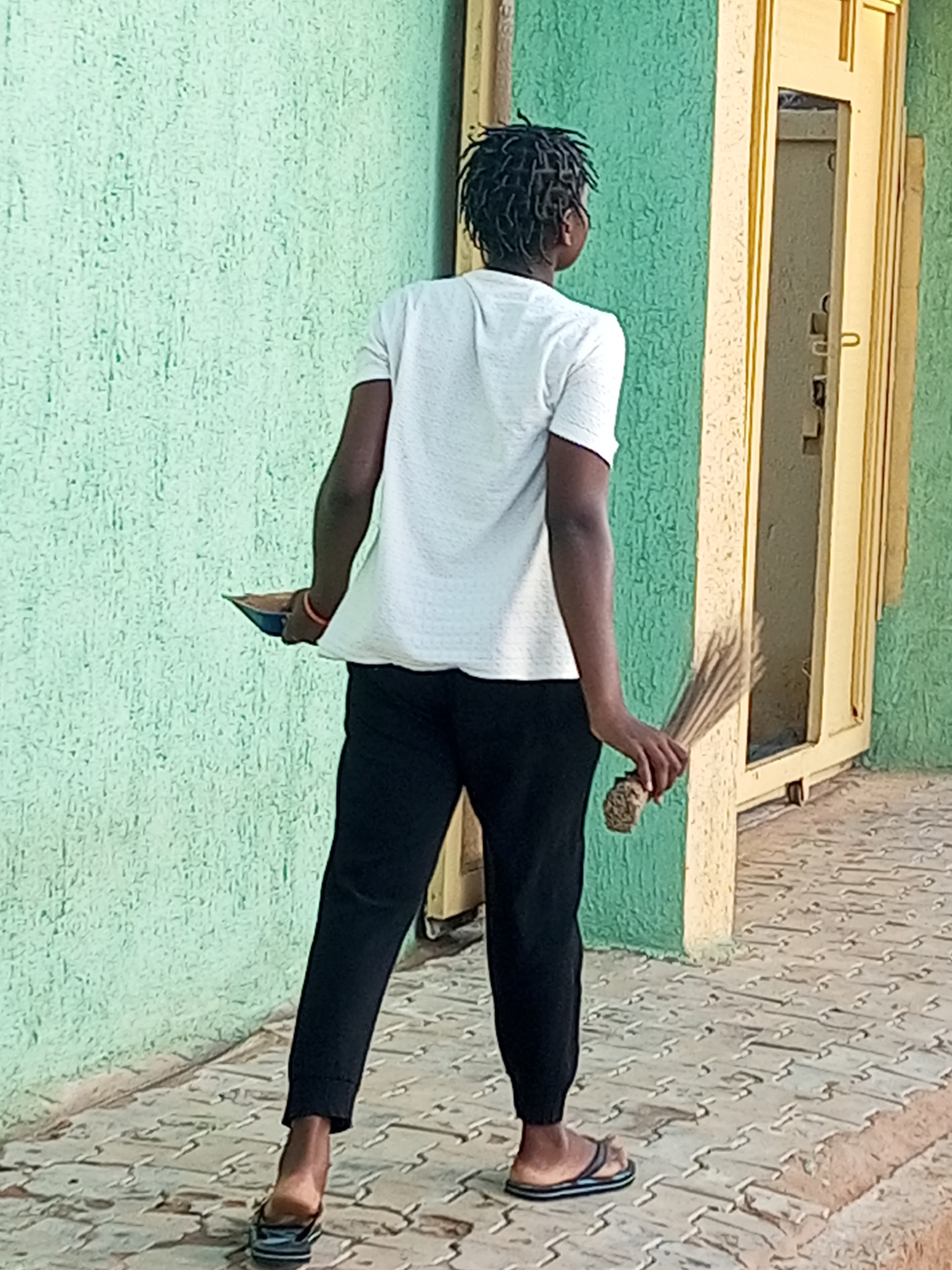 A person is standing outdoors in Gombe, Nigeria, facing away from the camera. They are wearing a white t-shirt and black pants. The person is holding a broom in their right hand and a dustpan in their left hand. They are wearing flip-flops. The background consists of a textured green wall and a yellow door. The ground is paved with interlocking bricks. The lighting suggests it is daytime.