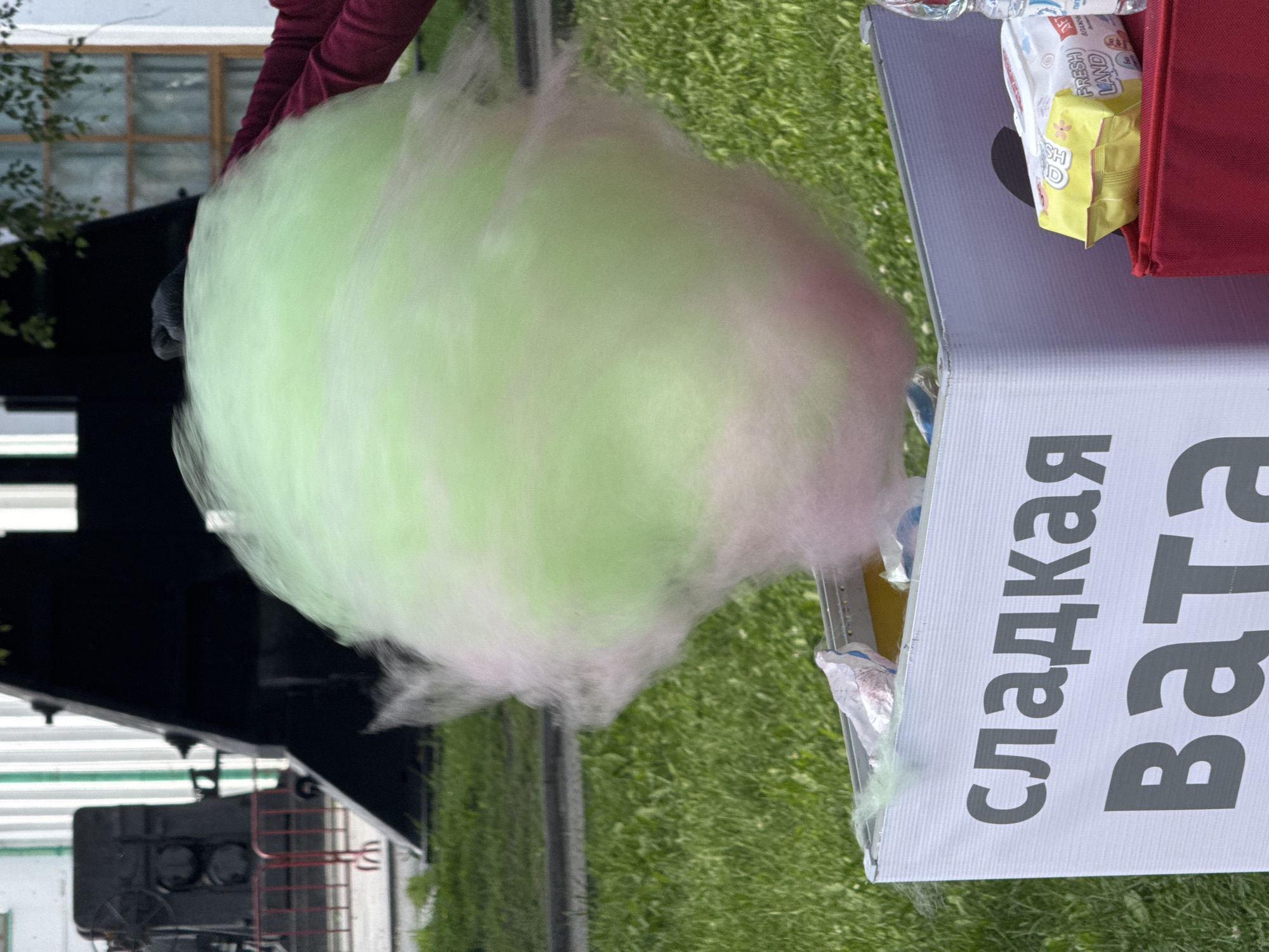 This media file captures an outdoor scene on a bright, possibly sunny day, given the strong lighting and shadows. The central subject is a large, fluffy ball of green and pink cotton candy, seemingly in motion or freshly made, with wisps of sugar extending outward. A person's hands, wearing burgundy sleeves, are partially visible, suggesting they are holding or preparing the cotton candy.
To the right of the cotton candy, a white table or stand is visible. On it, there are several items, including a red bag and a yellow and white package labeled "FRESH LAND" with a flower design, hinting at packaged snacks or ingredients. Below this, a larger white sign with the Cyrillic text "сладкая вата" (sweet cotton candy) is prominently displayed. The text is in large, gray, sans-serif font, with "ва" appearing to be the start of a second word. The setting appears to be a grassy area, possibly a park or an outdoor event space. In the background, a black structure with white horizontal lines and a red ladder-like element is visible, suggesting some sort of industrial or architectural feature, perhaps a train car. The overall impression is one of a festive, outdoor snack vendor at an event. The location context of Yekaterinburg, Russia, places this scene within a specific geographic and cultural setting.