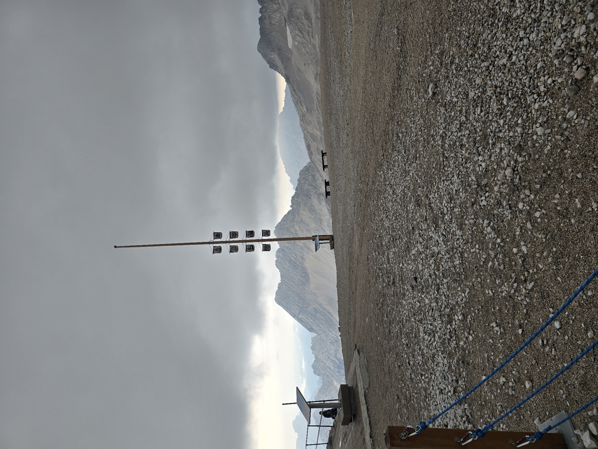 The image captures a panoramic view of a mountainous, rocky, outdoor location, likely a high-altitude viewpoint. In the foreground, a gravelly, uneven ground is visible, with a wooden post and blue ropes forming a safety barrier on the left side. Further back, a person is seated on a stone bench beneath a solar panel, near a metal railing.
Dominating the mid-ground is a tall wooden pole adorned with multiple small, square banners featuring emblems or crests. Two dark wooden benches are situated on the gravelly terrain to the right of the pole.
The background is a dramatic mountain landscape under a heavily overcast sky. Jagged, grey mountain peaks extend into the distance, with some snow visible on higher elevations. The overall atmosphere is muted and somewhat somber due to the cloudy weather.
Based on the presence of such a high-altitude viewpoint with distinct banners and the geological formations, this scene is highly likely to be the Zugspitze, Germany's highest peak, located near Garmisch-Partenkirchen. The banners might represent regional symbols or historical affiliations. The benches and solar panel suggest a managed tourist area.