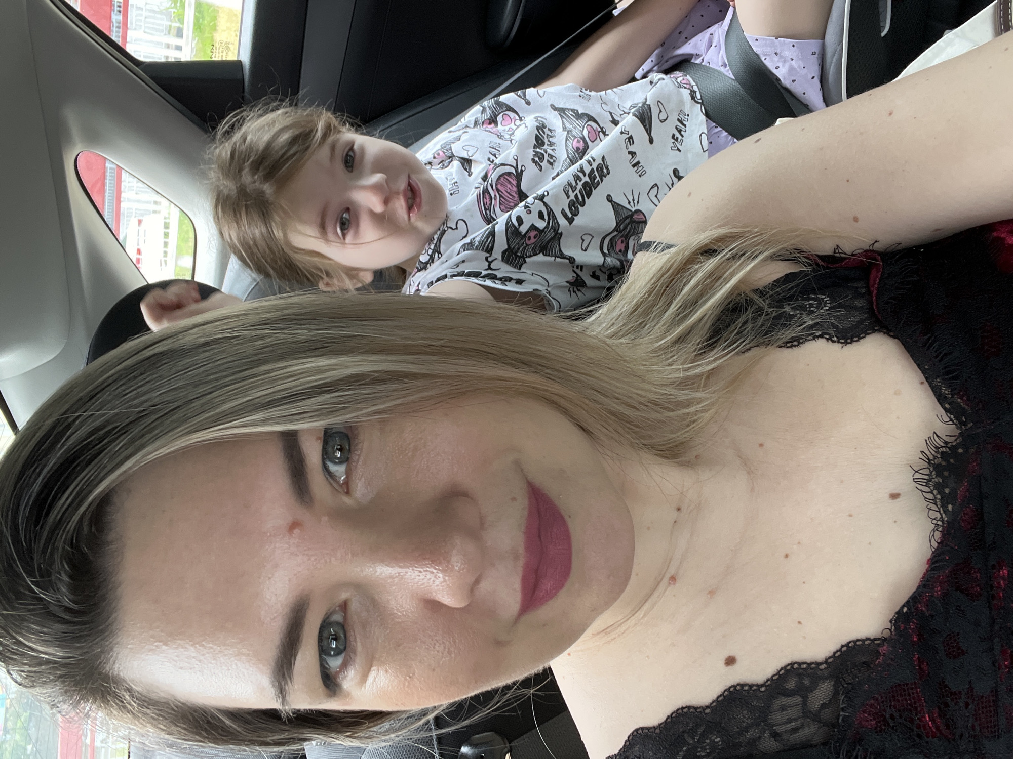 In the backseat of a vehicle, a woman is taking a selfie with a young girl seated behind her. The woman, positioned in the foreground on the left, has light brown hair and is wearing a black lace top. She is looking directly at the camera with makeup on. The girl, in the background on the right, has blonde hair and is wearing a white t-shirt with a cartoon print and purple shorts. She is also looking towards the camera with a slightly open mouth. The interior of the car is visible, including the seatbelts and the headrest of the driver's seat. The window behind the girl shows a blurry view of a red and white structure, suggesting an outdoor environment. The image does not contain any information that places it in Sochi, Russia.