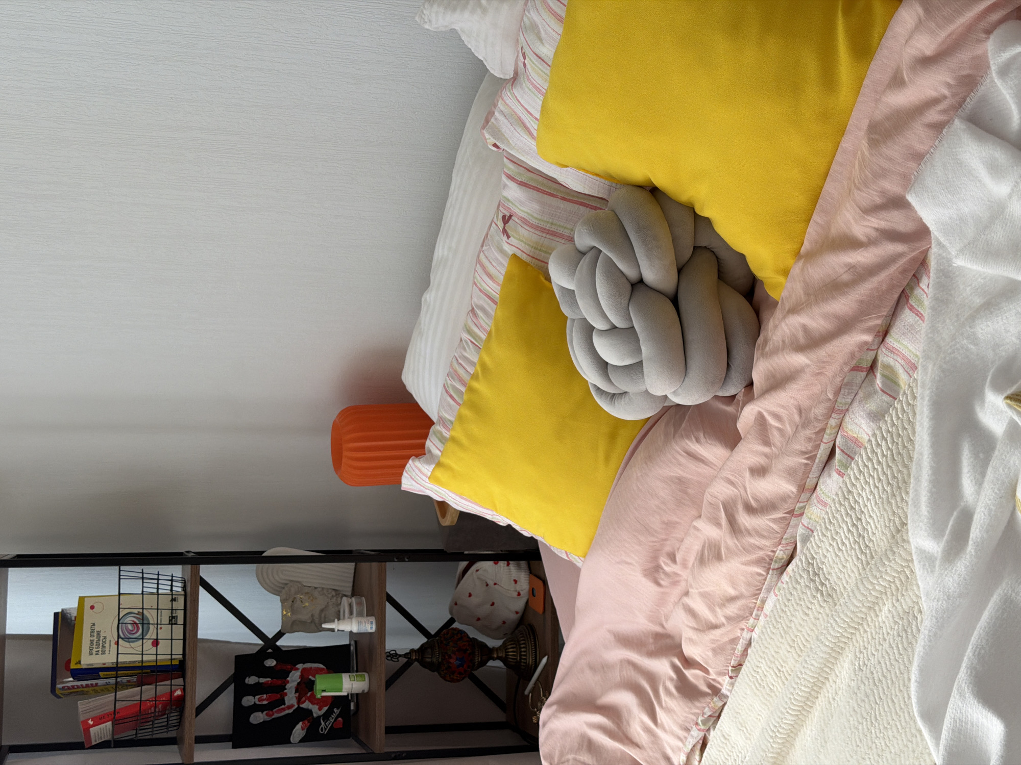 A bedroom scene shows a neatly made bed with pink and white striped bedding, a light pink duvet, and several pillows. Two bright yellow square pillows are prominently displayed, along with a grey knotted pillow. A white textured blanket is folded at the foot of the bed, and another white blanket is partially visible. To the left, a dark metal shelf unit contains books, a framed handprint artwork, a white bust, a decorative lamp, and a small orange object. An orange cylindrical lamp is visible on the right side of the bed. The room has a plain white textured wall. The overall impression is one of a clean and organized personal space.