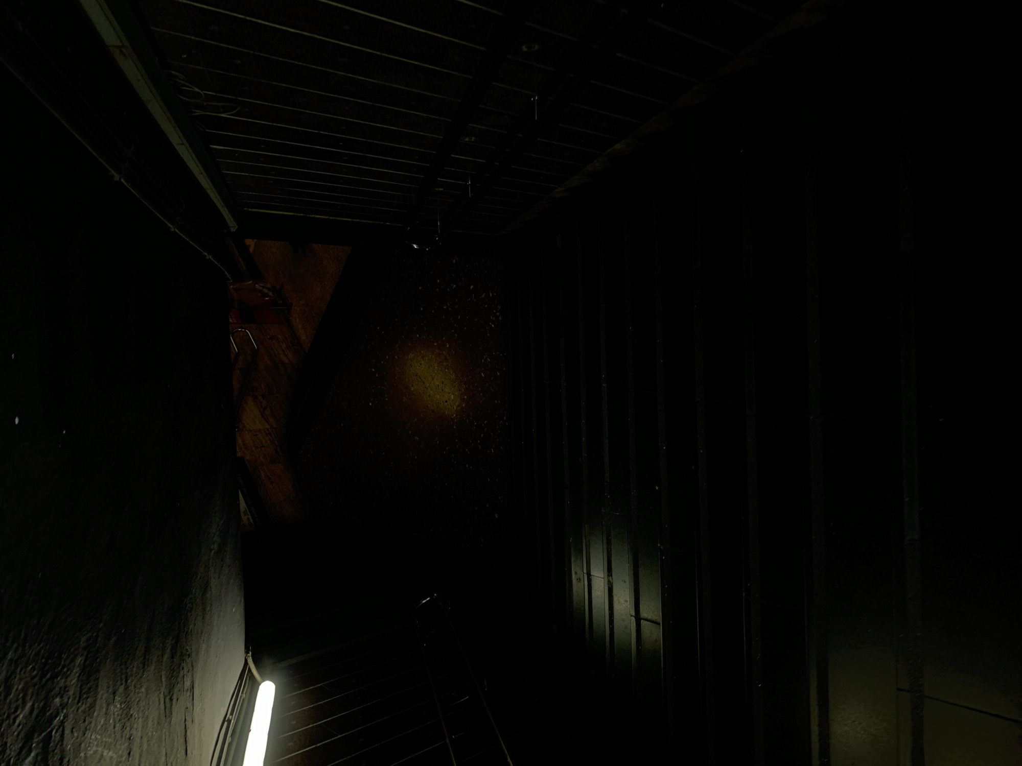 The image shows a dimly lit underground tunnel in Barcelona, Spain. The ceiling is constructed of parallel metal beams. A light source illuminates a section of the wall in the center of the image. A vertical light fixture is visible on the left side. The floor appears to be made of metal grating. The walls are dark and rough.