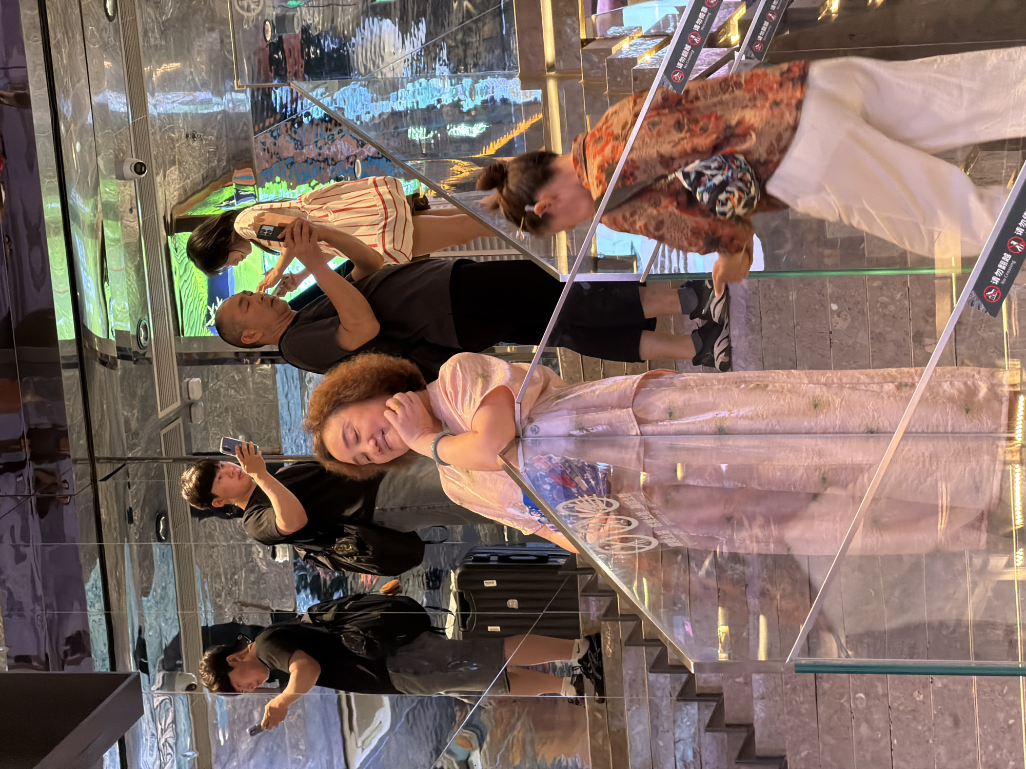 In the Yuzhong District of China, a group of individuals are seen on a multi-level staircase with a reflective ceiling. In the foreground, a woman in a pink dress stands facing left with her eyes closed and hand touching her face, a small patterned bag hanging from her shoulder. To her right and slightly behind her, another woman in a rust-colored patterned top and white pants is walking up the stairs, also with a patterned bag. On the stairs above and behind the woman in pink, two men are visible. One man in a black t-shirt and shorts holds up a smartphone, looking at it. Another man, also in a black t-shirt and shorts, is further up the stairs, also holding a smartphone. In the background, a young woman in a white and red striped dress stands near a man in a black t-shirt. The environment features marble-like stairs and a highly reflective, chrome-like ceiling that mirrors the scene, creating a distorted and busy visual effect. Security cameras are visible on the ceiling. Several "No Crossing" signs are posted along the railing of the stairs.