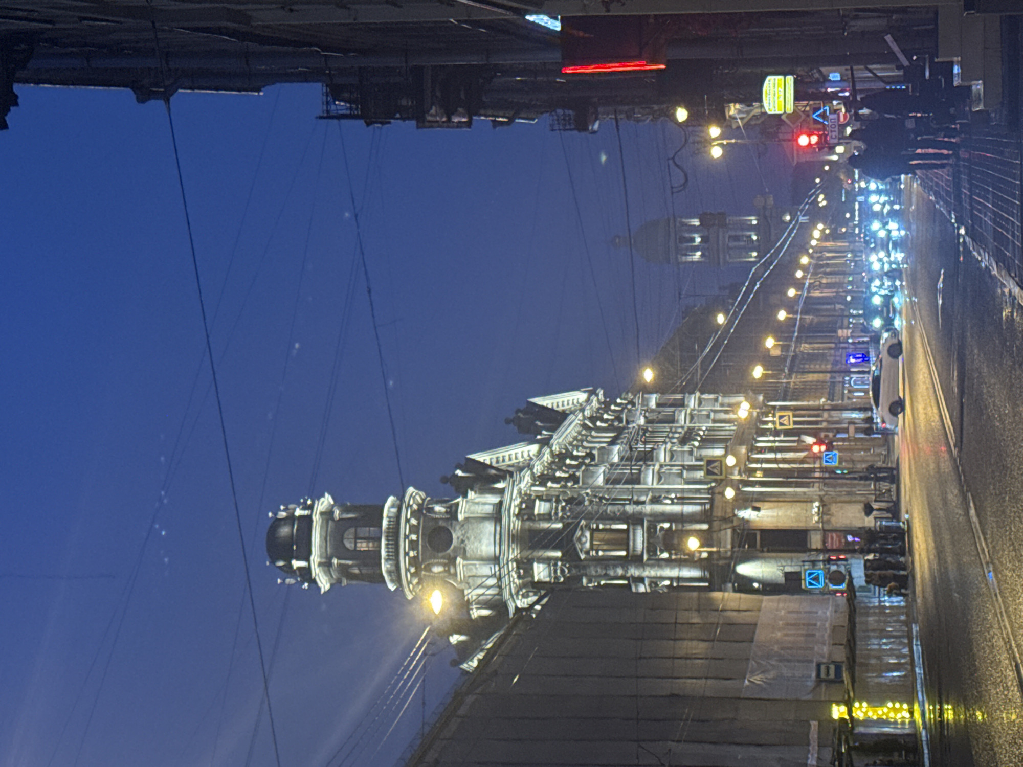 This nighttime shot of Saint Petersburg, Russia features the Anichkov Bridge over the Fontanka River. On the left, the Stroganov Palace is visible. On the right side of the image, a wet street runs into the distance, lined with streetlights and buildings. Cars are on the road, and traffic lights are visible. People are walking on the sidewalk. Power lines stretch across the sky.