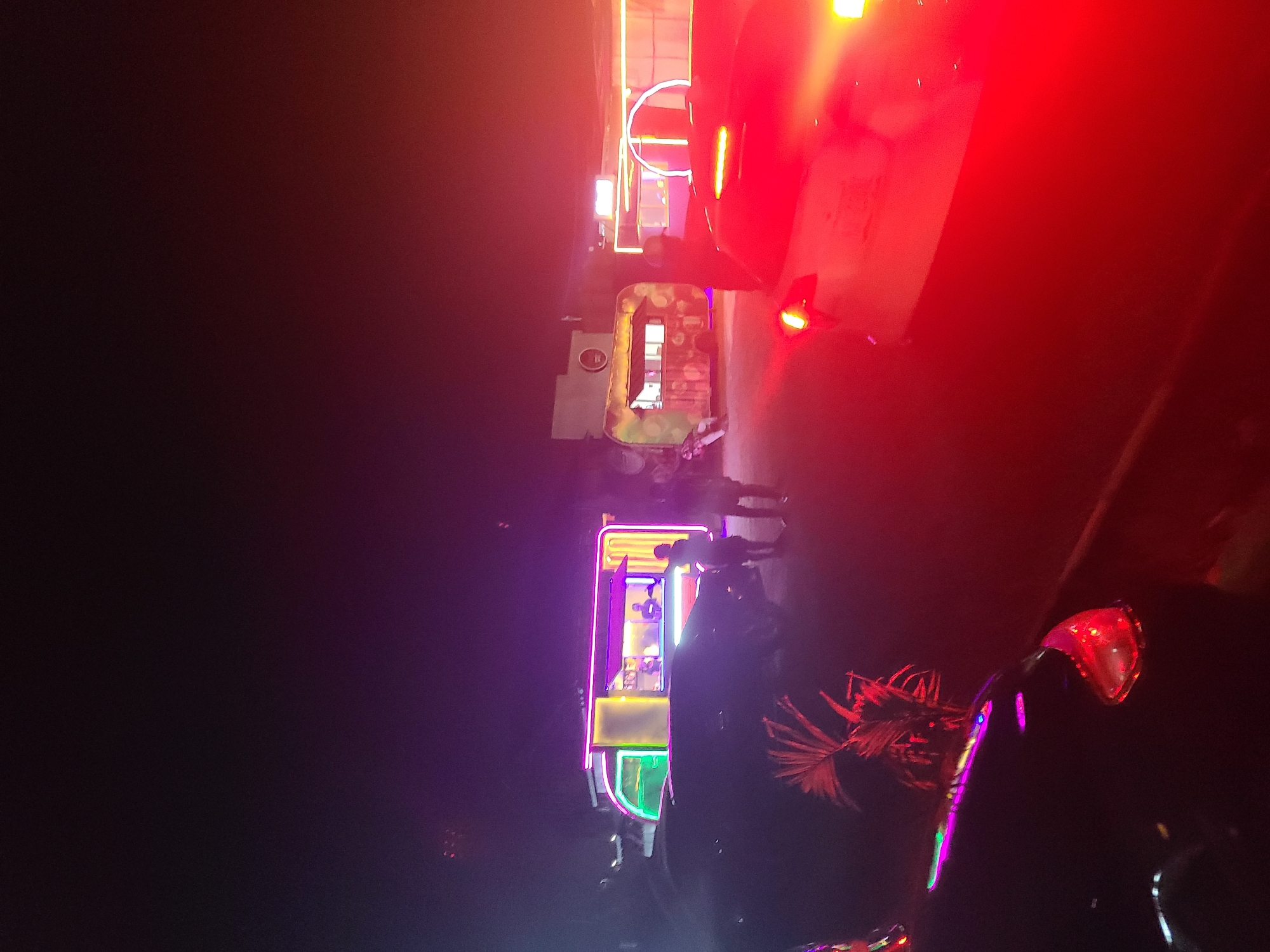 At night in Jos, Nigeria, the image depicts an outdoor scene with bright neon lights emanating from a building. A structure stands out with multiple floors, each outlined with a different color of neon light (purple, green, yellow). A partial view of a vehicle, possibly a car or truck, is visible in the frame. The image is overexposed in some parts due to the bright lights. Some people are present, although silhouetted or indistinct due to the lighting conditions.