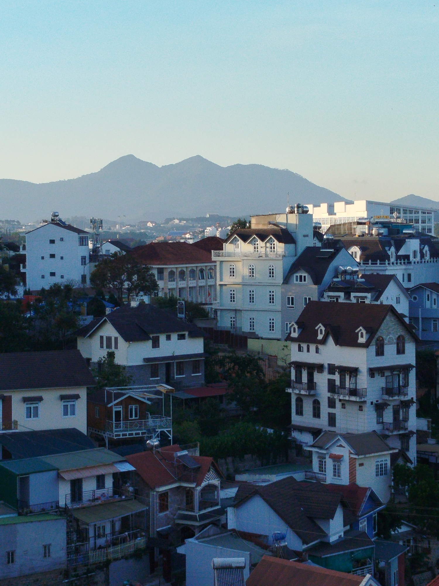 The photograph presents a daytime, elevated view of a section of Da Lat, Vietnam, with mountains in the background under a light blue sky. The foreground consists of closely packed buildings, varying in height and architectural style. Many feature pitched roofs, and their facades display a mix of white and colored paint. Trees and greenery are interspersed among the buildings. In the background, there are two mountains with a hazy appearance due to distance or atmospheric conditions. The lighting suggests either early morning or late afternoon.