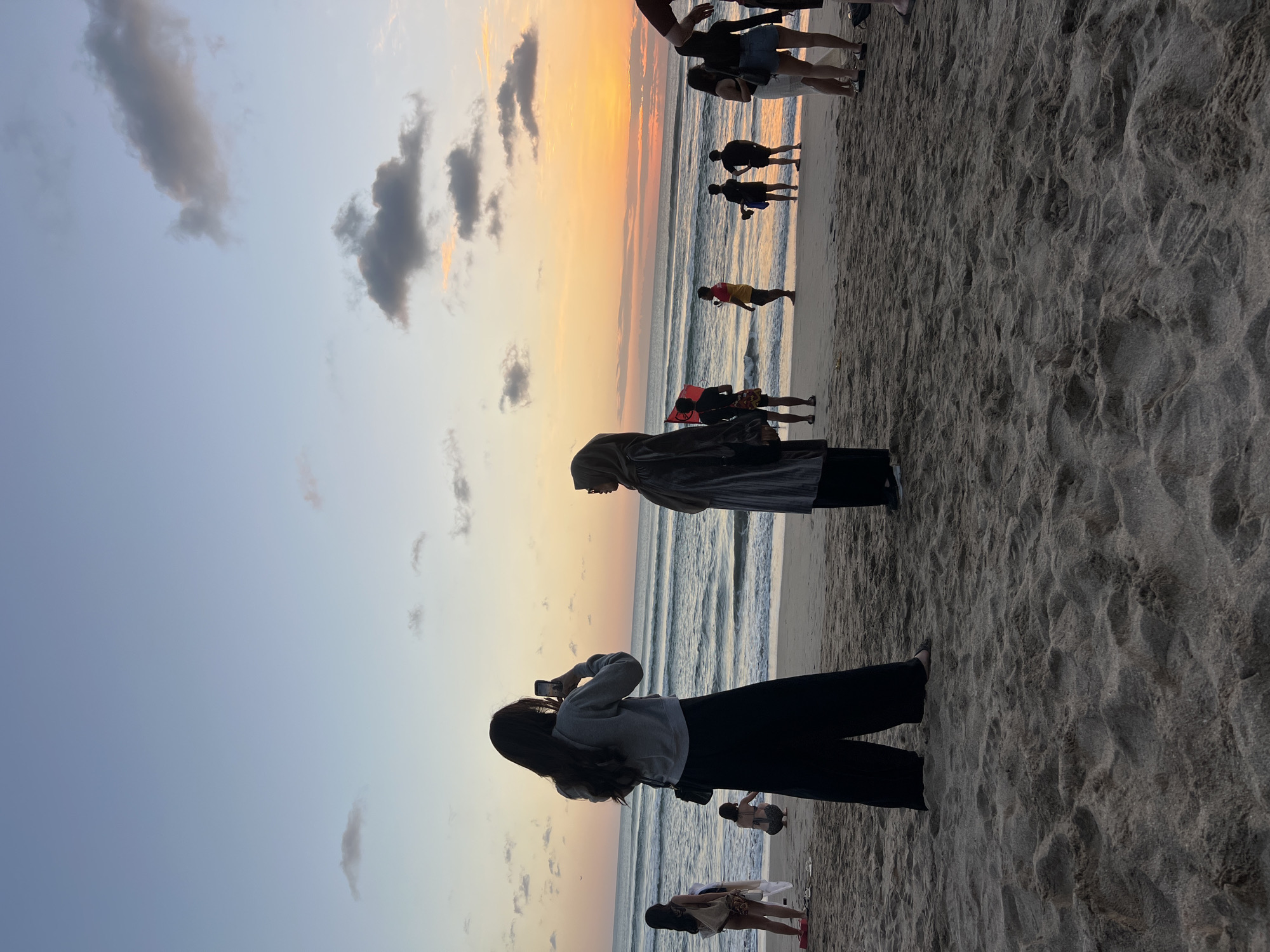 In Kuta, Indonesia, a group of people are gathered on a sandy beach during sunset. The sky is a gradient of blue and orange, with scattered clouds. The ocean has moderate waves. In the foreground, a woman with long dark hair is taking a photo with her phone, wearing a gray hoodie and black pants. To her right, a woman in a hijab and a striped dress stands facing the ocean. Further back, several other individuals are scattered on the beach, some standing and some crouching. The sand is light brown and textured, with footprints visible. The overall atmosphere suggests a relaxed evening gathering by the sea.