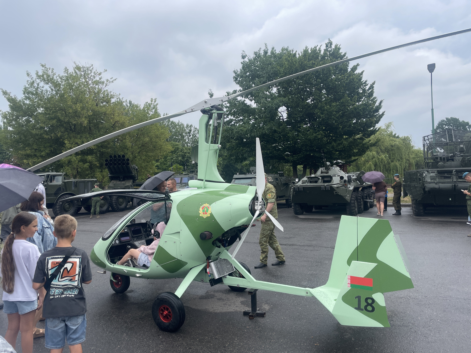 This image captures an outdoor event, likely a public exhibition or festival, in Brest, Belarus, on a cloudy day. The central focus is a small, two-seater autogyro painted in a camouflage pattern of light and dark green. The autogyro has a prominent rotor on top and a propeller at the front. A young girl, wearing a pink jacket and light blue shorts, is seated in the cockpit, appearing to be a passenger. Beside the autogyro, a man in a military uniform stands, possibly an attendant or presenter.
In the background, several military vehicles are visible. To the left, a dark green military truck with multiple wheels is parked. Behind the autogyro and to its right, two armored personnel carriers are positioned. People are scattered throughout the scene, some holding umbrellas, indicating light rain or overcast conditions. Several trees with dense green foliage line the perimeter of the event space. The ground is paved and wet, suggesting recent precipitation. A flag of Belarus is prominently displayed on the tail of the autogyro, along with the number '18'. The overall atmosphere appears to be one of public interest and engagement with military equipment.