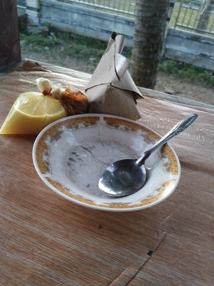 This image, captured in Jambo Dalam, Indonesia, displays a static scene of a meal setup on a wooden table. The foreground consists of a light brown wooden surface with prominent grain. Centrally positioned is an oval white ceramic plate featuring a gold and white patterned border; the plate's interior exhibits dried residue. A silver-colored metal spoon rests within the plate. To the left of the plate are two small packages: one is a clear plastic bag containing a translucent yellow liquid, secured with a red tie; the other is a smaller, irregularly shaped item wrapped in brown paper and tied. Behind these rests a larger, triangularly folded package made of brown paper. The background, out of focus, reveals an outdoor environment featuring a dark tree trunk on the right and a grey metal fence or railing visible through green foliage. No individuals are present.