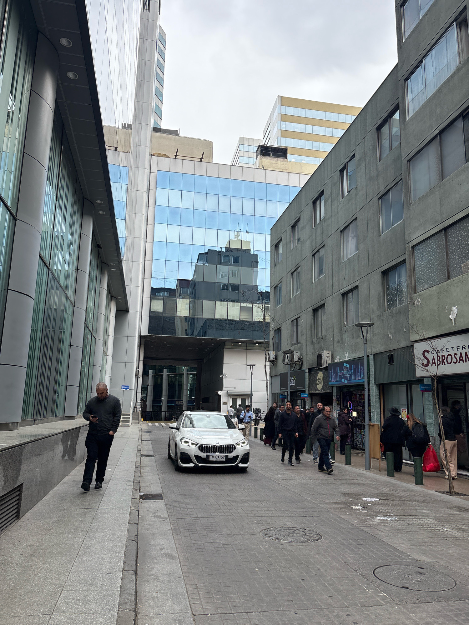 The image depicts a street scene in Santiago, Chile, with modern architecture dominating the urban landscape. On the left, a tall building with a glass facade and metallic accents reflects the overcast sky. A man in a dark sweater and black pants is walking on the sidewalk, looking at his phone. The sidewalk is paved with light-colored tiles.
The street itself is paved with grey brickwork and is relatively narrow. A white BMW sedan with license plate "TV CX-57" is driving down the street. On the right side of the street, a darker, older building with multiple windows rises several stories. Various shops and businesses are visible at street level, including "CAFETERIA SABROSAN." Several pedestrians are walking along the sidewalk on the right.
In the background, other tall buildings, some with glass facades and others with lighter-colored exteriors, are visible, contributing to the dense urban environment. The sky is cloudy, suggesting a temperate climate. The overall impression is one of a busy, modern city with a mix of contemporary and older architectural styles.