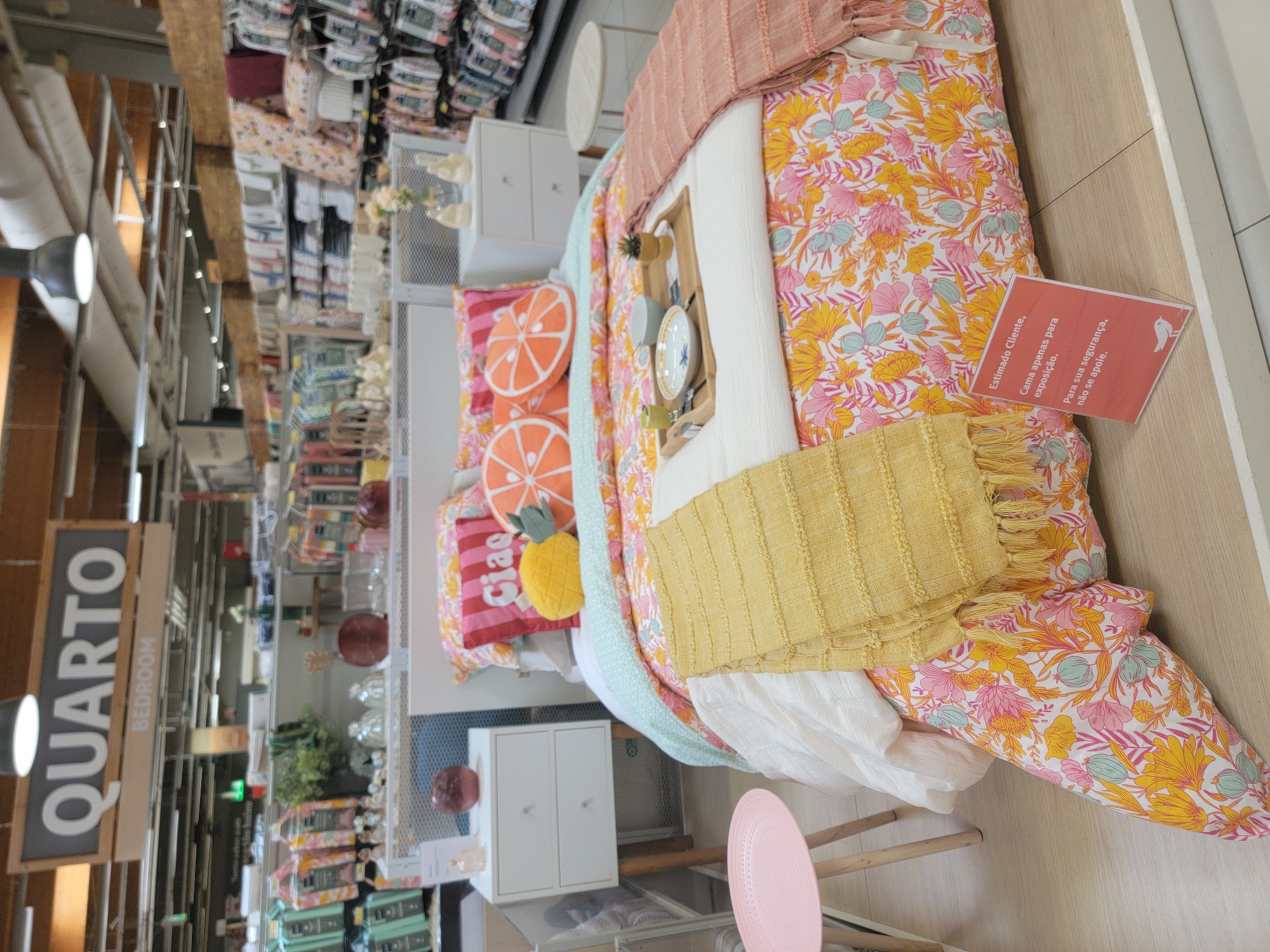 The image shows a bedroom display in a store, likely a furniture or home goods store. A bed is set up with a colorful floral duvet cover, several decorative pillows including one shaped like a pineapple and two shaped like orange slices, and a yellow textured throw blanket. A wooden tray with a bowl, plate, and a small plant sits on the bed. There are two white nightstands with wooden legs on either side of the bed. A small pink stool is visible to the left of the bed. In the background, shelves are stocked with various home goods, including bedding and decorative items. A sign at the top of the display reads "QUARTO" and "BEDROOM." A small orange sign with white text is propped up on the bed, stating "Estimado Cliente, Cama apenas para exposição. Para sua segurança, não se apoie." (Dear Customer, Bed for display only. For your safety, do not lean on it.). The lighting suggests an indoor retail environment. The mention of Faro, Portugal in the prompt indicates this store is located there.