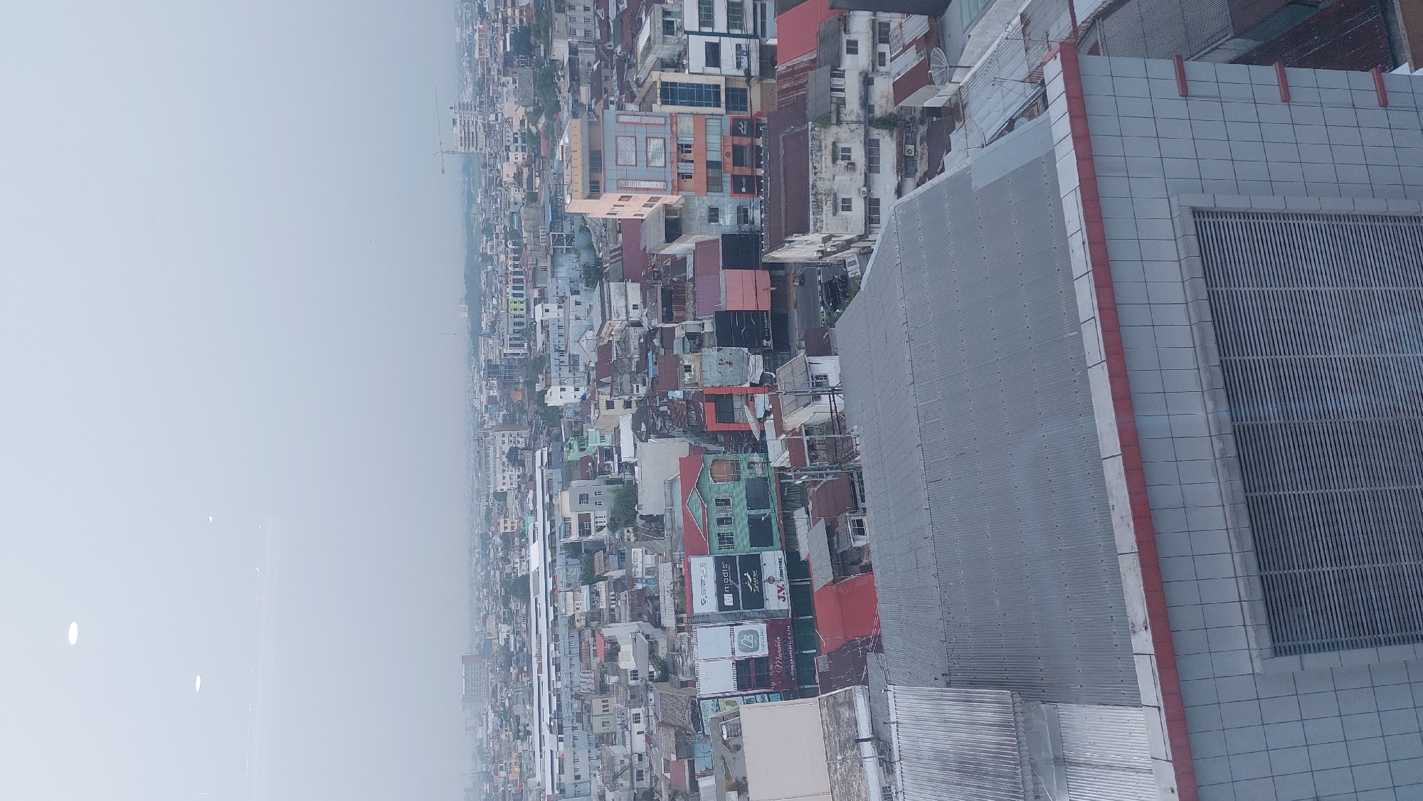 This is an aerial, high-angle shot of a dense urban environment in the City of Medan, Indonesia, likely taken during daytime given the ambient light. The foreground is dominated by the rooftop and upper facade of a modern building. The rooftop is made of corrugated metal, with some lighter-colored sections and a red trim running along the edge of the visible facade. The facade itself is clad in light-colored tiles with visible grout lines and features a large, louvered vent or window.
Beyond this foreground structure, the image reveals a sprawling cityscape composed of numerous buildings of varying heights and architectural styles, packed tightly together. The buildings are predominantly residential or commercial, with many featuring pitched roofs, some made of corrugated metal and others of tiles in red or brown hues. Some buildings display signs, indicating commercial establishments.
The color palette of the cityscape is muted, dominated by grays, whites, browns, and some patches of green from trees peeking through. The overall atmosphere suggests a hazy or overcast sky, which softens the details and contributes to a slightly diffused quality of light across the scene. There are no visible people or distinct vehicles in the image, focusing the view solely on the built environment and its density. The scene conveys a sense of urban sprawl and the continuous, interconnected nature of city development.
