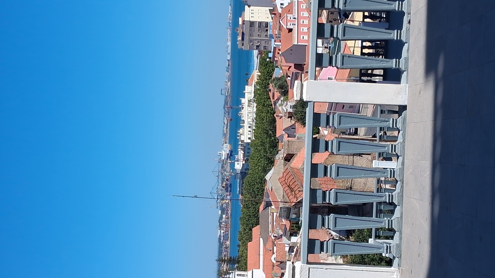 A high-angle, full shot captures a sunlit, panoramic view of Setúbal, Portugal. In the foreground, a blue-grey decorative balustrade with ornate posts spans the lower portion of the image, casting a shadow on the paved terrace below. Beyond the balustrade, a dense collection of terracotta-roofed buildings, typical of Mediterranean architecture, stretches across the mid-ground. A white steeple, likely belonging to a church, is visible on the far left. The background is dominated by a wide, blue expanse of water, likely the Sado River estuary, with a busy port in the distance. Numerous ships, including cargo vessels and a large cruise ship, are moored or navigating the water, alongside prominent industrial cranes and structures that define the port's activity. A lush green line of trees separates the residential area from the waterfront, adding a natural element to the urban landscape. The sky above is a clear, vibrant blue, devoid of clouds.