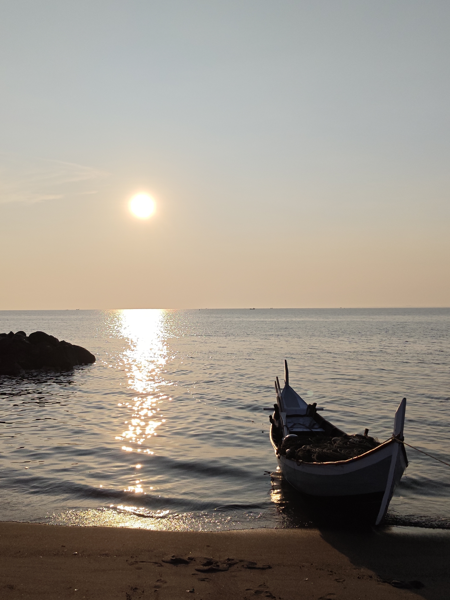 The photograph captures a tranquil seaside scene in Lhokseumawe, Indonesia. A traditional fishing boat rests on the sandy shore at low tide. In the background, the sun hangs low, casting a golden reflection across the still water. A small cluster of rocks is visible to the left, partially submerged in the sea. The sky is clear, with a gradient of light blue to yellow near the horizon. Several small boats are distantly visible on the horizon. The boat on the shore is loaded with fishing nets. There are no people visible in the scene.