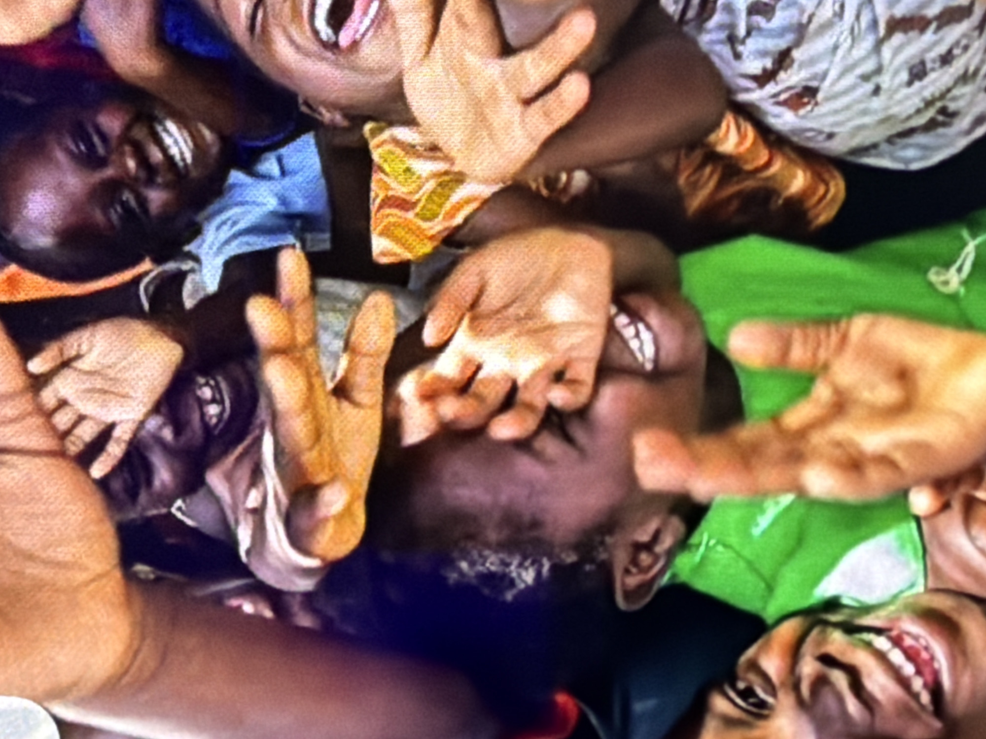 A close-up shot captures a group of children and young adults in what appears to be an outdoor setting, though the specific location in the city of Unknown, Unknown is not identifiable from the image. The subjects are primarily of African descent, with varying skin tones and hair textures. Many of them are smiling, laughing, and looking directly at the camera, suggesting a candid and joyful moment.
In the foreground, a child in a bright green shirt is prominent, their face partially obscured by hands. Their mouth is wide open in a laugh, and their eyes are crinkled with mirth. To the right, another individual, likely an adult due to their larger hands, is also laughing, their mouth open and tongue visible.
Other faces are visible in the background, some peeking through gaps in the hands. One child in the upper center is wearing a blue shirt and has a broad, toothy grin. The overall impression is one of togetherness and shared happiness. The lighting suggests it is daytime, and the focus is sharp on the faces, with a slight blur in the background, indicating a shallow depth of field. The vibrant colors of the clothing, particularly the green shirt and an orange and yellow patterned garment, add to the lively atmosphere of the scene.