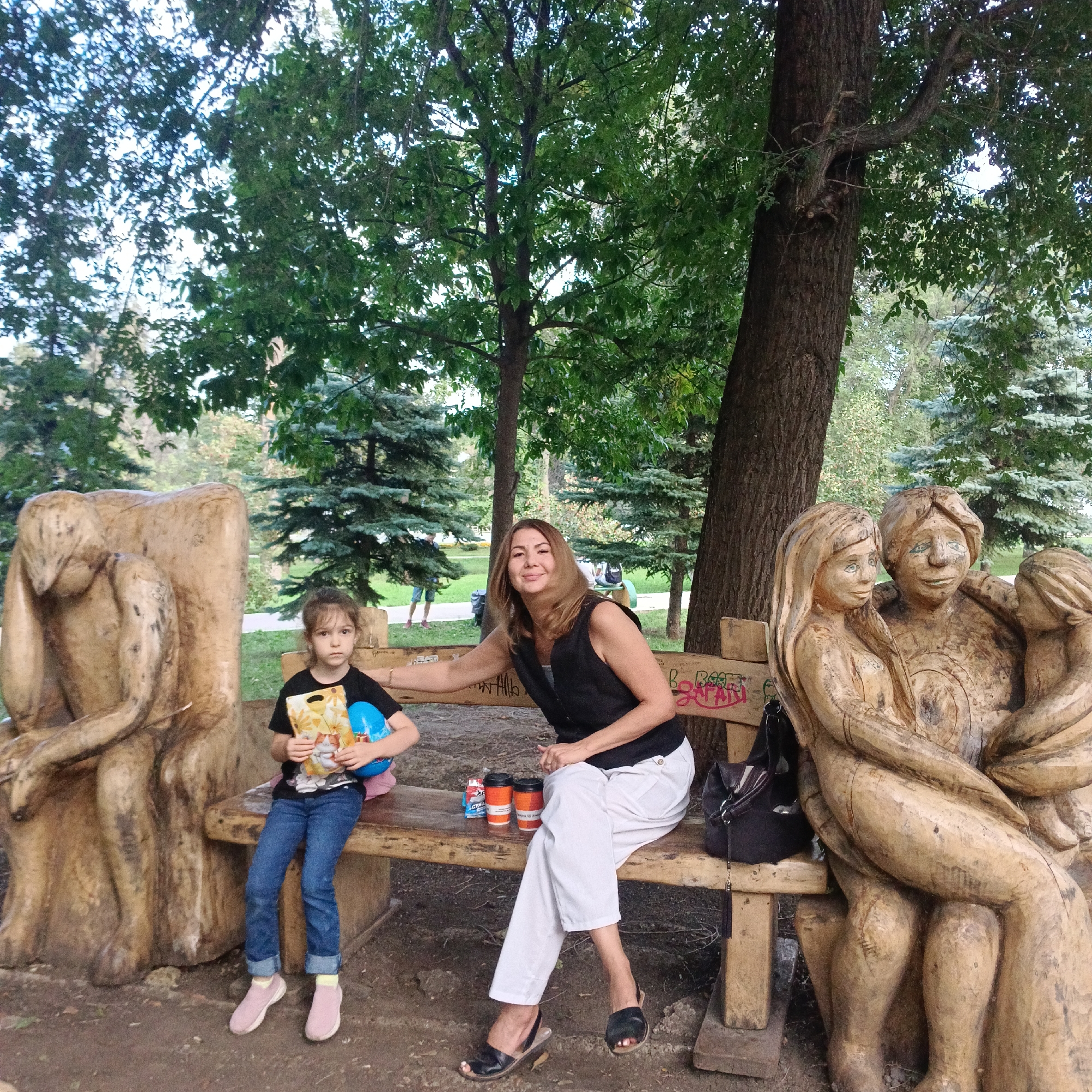 In a park in Samara, Russia, a woman and a young girl sit on a carved wooden bench. The woman, with shoulder-length blonde hair and wearing a black vest over a white shirt and white pants, smiles at the camera. Her left arm is extended towards the girl. The girl, with brown hair pulled back, wears a black t-shirt and blue jeans. She holds a book with a yellow cover and a blue toy. Two orange coffee cups and a small package are on the bench between them. The bench itself is carved from wood, with figures of people on either end. A dark bag hangs from the back of the bench. The background features large trees, greenery, and a pathway. The ground is covered with dirt and fallen leaves.