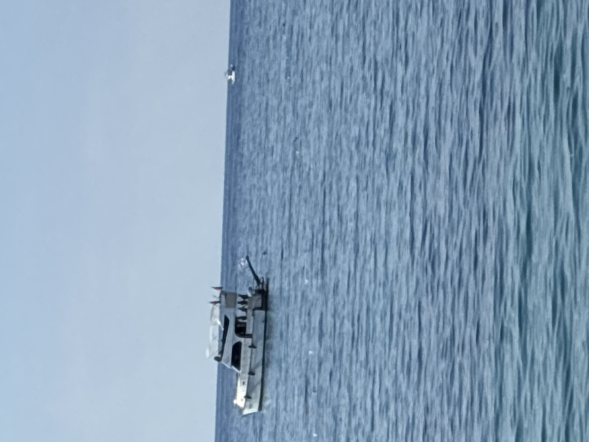 A white motor yacht is anchored in the deep blue waters of the Mediterranean Sea, near Alanya, Turkey. The yacht has a two-level cabin, a white canopy, and several flags on its stern. There are several people visible on the deck of the yacht. In the distance, a smaller white boat is also visible on the horizon. The sky is clear and pale blue. The water is calm with slight ripples.