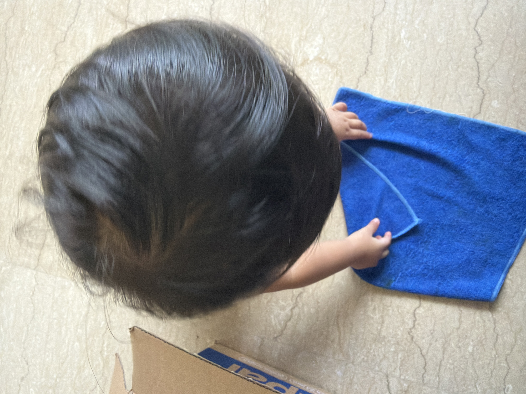 The overhead shot captures a toddler's head and a hand as they interact with a blue microfiber cloth on a marble-patterned floor. The child, viewed from above, has dark hair. Their right hand is visible, gently touching the cloth, which is folded in half and lies on the floor. To the left of the child's head, a portion of a cardboard box is visible, with blue tape along its edge. The scene is indoors, on a light-colored, possibly travertine or marble, tiled floor with faint dark veins. The lighting appears to be natural, coming from above, casting soft shadows. The image does not provide any specific details to identify the location as Central Jakarta, Indonesia.