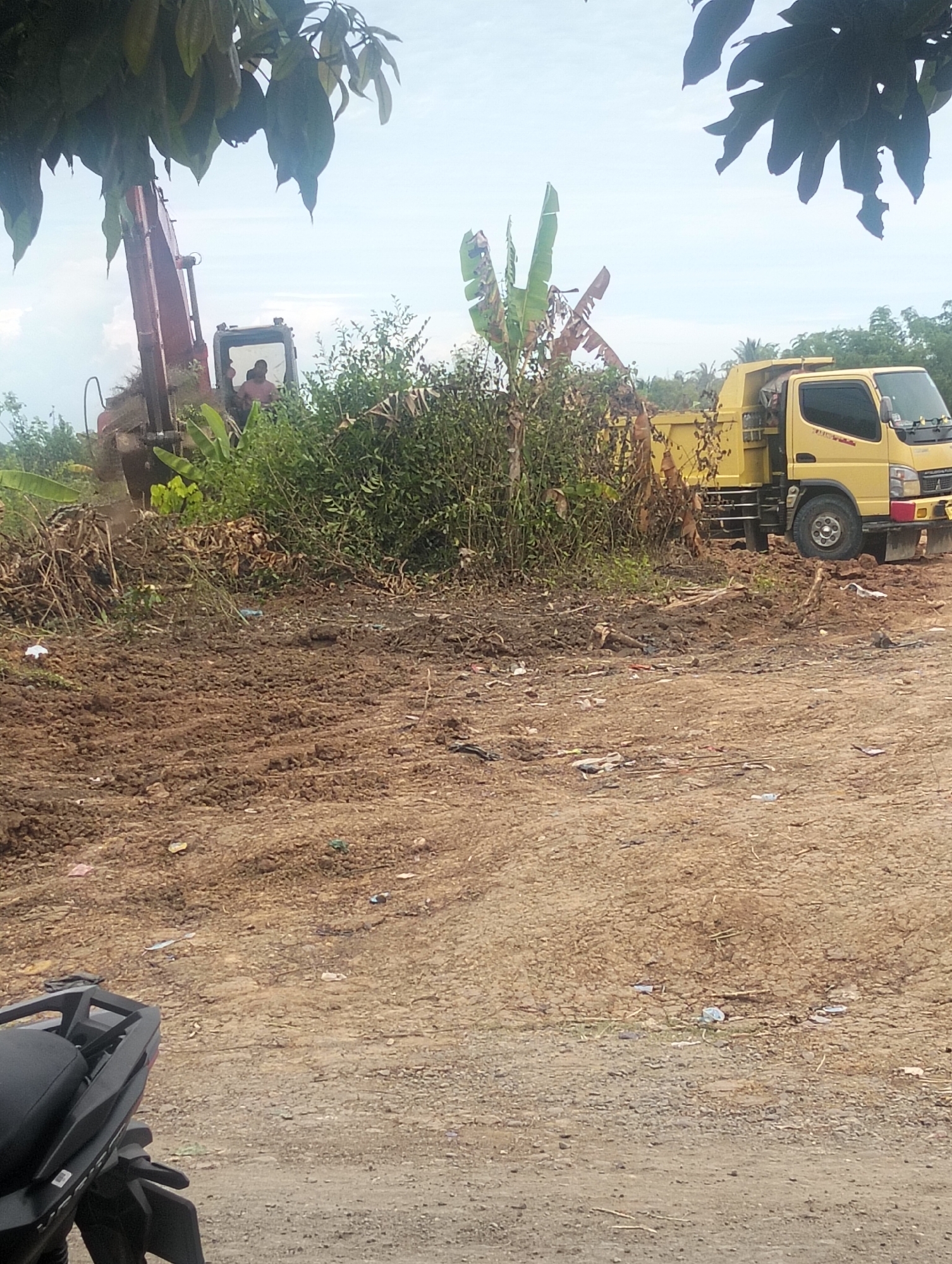 In Payagaboh, Indonesia, an excavator is actively working, with its arm extended and bucket lowered, near a yellow dump truck. The ground is uneven and appears to be dirt, with some vegetation and debris scattered around. The sky is cloudy. A portion of a black motorcycle is visible in the foreground, partially obscuring the lower left quadrant of the image.