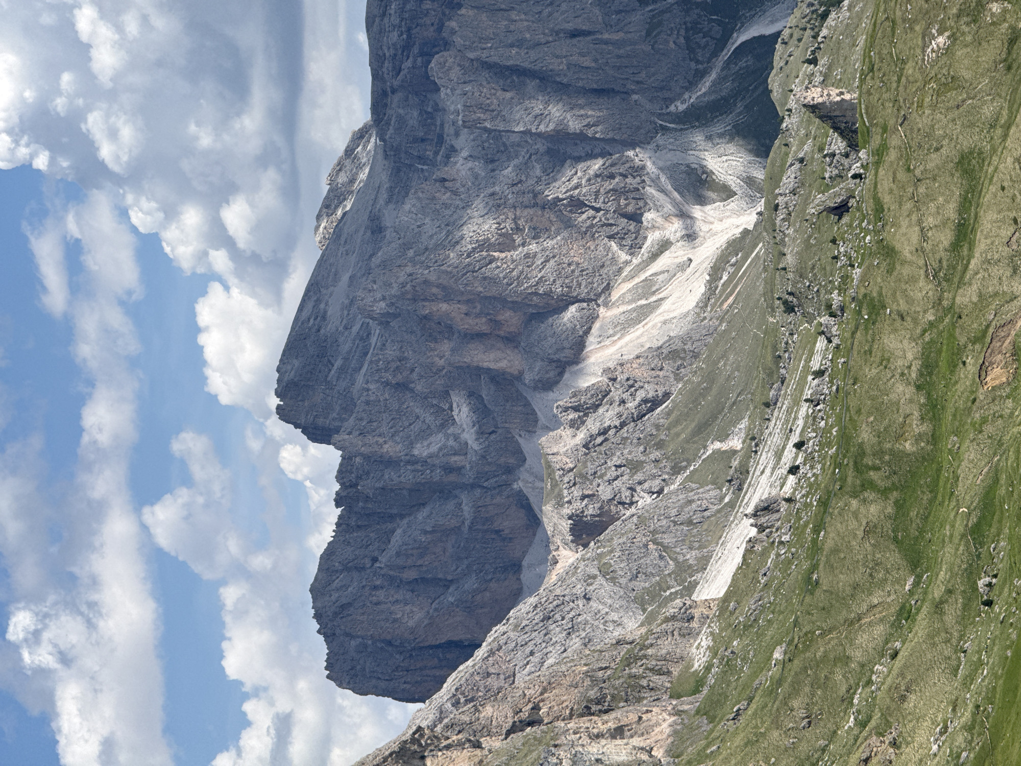 This media file is an image of a steep, rocky mountain range under a partly cloudy sky. The dominant subjects are the natural landscape itself, with a focus on the rugged texture of the rock faces and the contrast between the barren rock and the green, grassy slopes. There are no visible people or man-made objects, suggesting a remote and natural setting.
The image captures a daytime scene, with bright sunlight casting shadows that highlight the contours of the mountains. The sky is a mix of blue and white clouds, indicating fair weather with some cloud cover. The location appears to be a mountainous region, likely in the Alps, given the type of terrain and vegetation. The specific location context of Santa Cristina Gherdëina suggests the Dolomites, known for their dramatic rock formations.
Notable details include the stark, jagged edges of the rock cliffs, some patches of snow or scree on the mountainside, and the lush green grass that covers the lower slopes. The sheer scale of the mountains is emphasized by the absence of any reference points for size. The image conveys a sense of grandeur and the raw power of nature. There is no visible text in the image.
