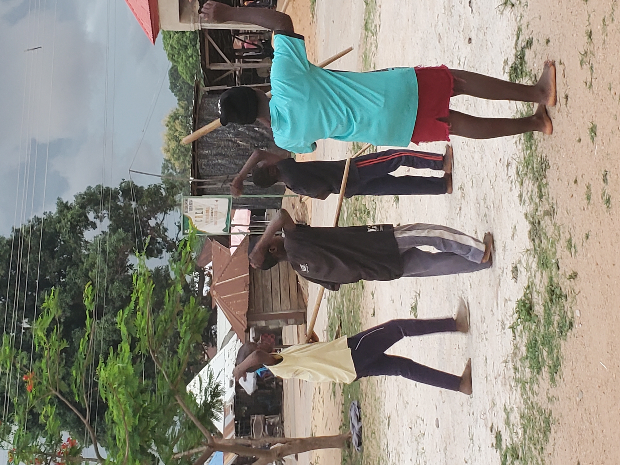 In Mararaba, Nigeria, four barefoot individuals are lined up performing an exercise outdoors. Each person holds a stick across their shoulders, behind their necks. The first person is wearing a light blue shirt and red shorts. The second person wears a dark shirt with red stripes down the pants. The third person wears a dark shirt and gray pants with a white stripe. The fourth person wears a yellow shirt and dark pants. There is a tree and some buildings visible. The sky is overcast.