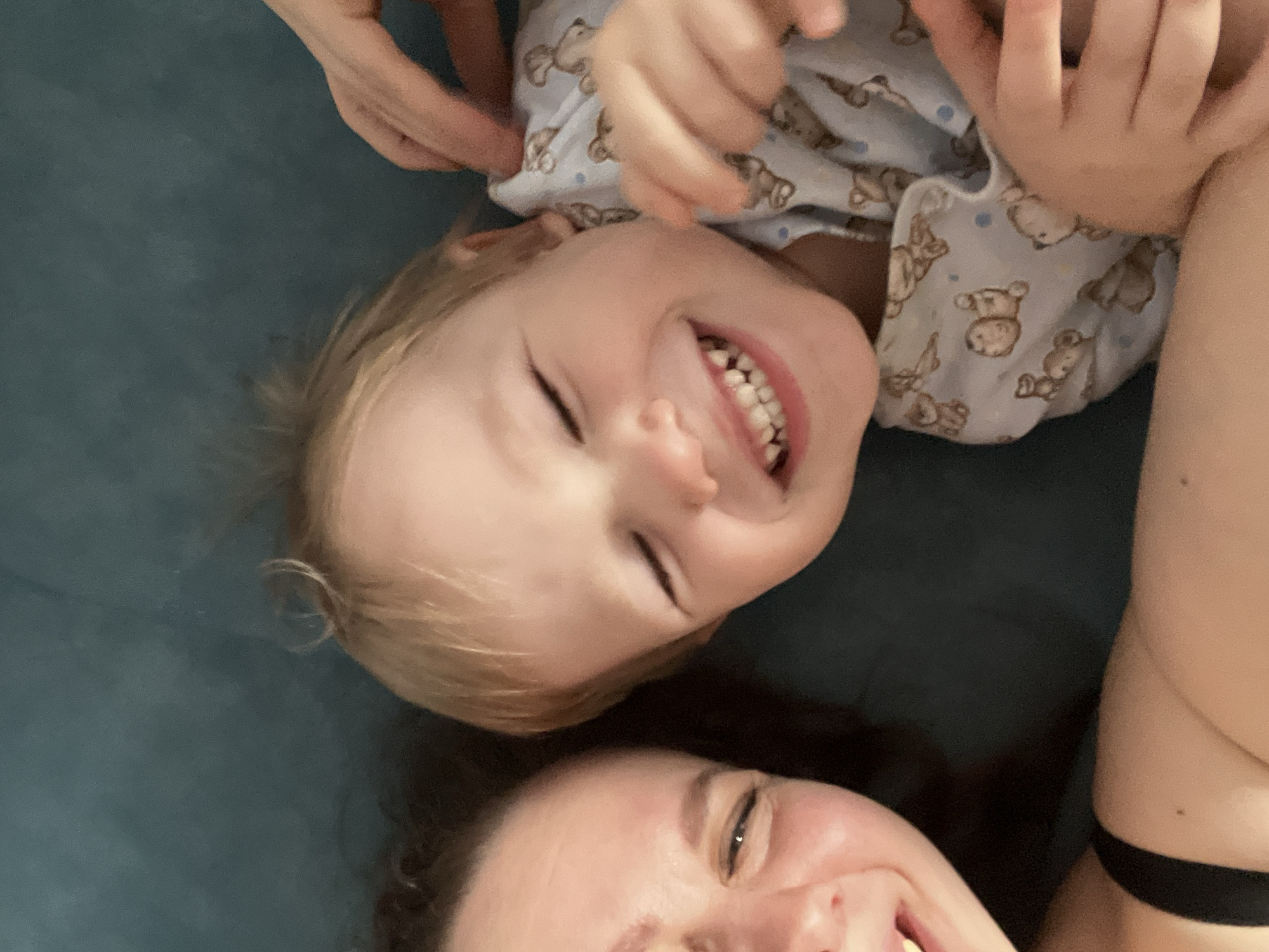 A close-up, slightly angled shot shows a woman and a child lying on what appears to be a teal surface. The child, wearing a light blue pajama top patterned with bears, is in the upper portion of the frame, smiling with closed eyes. The woman's face is in the lower part of the frame, also smiling. Part of a hand is visible near the child's head. The city of Chaikovsky, Russia is not discernible in the image.